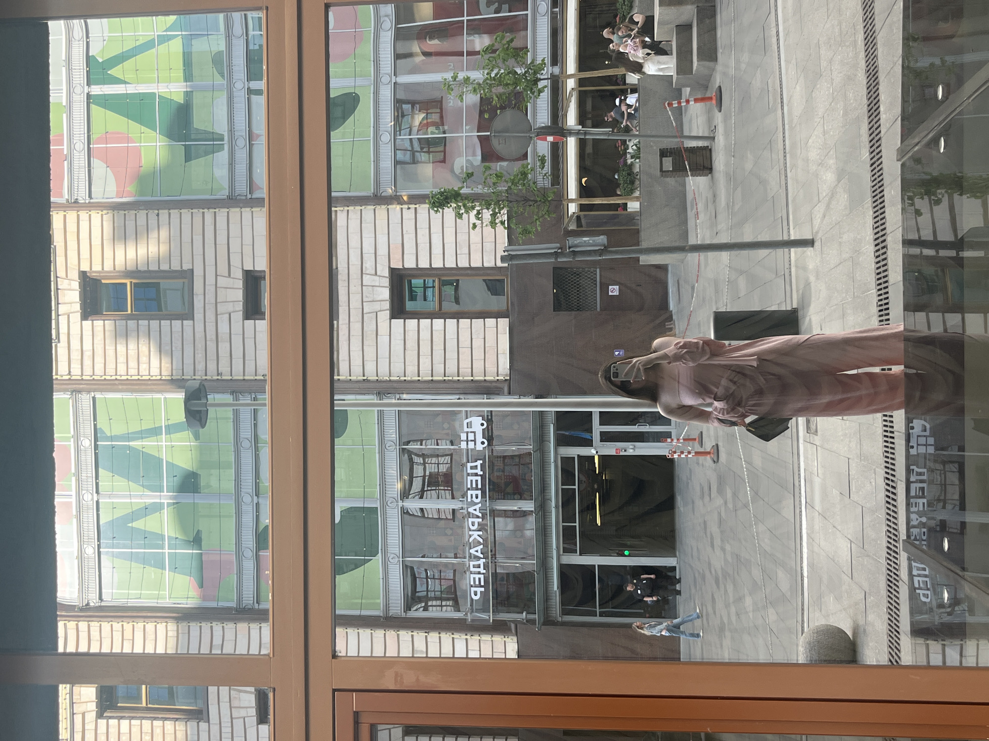 The image shows a view through a window, likely in Moscow, Russia. Reflections are visible on the glass, including the reflection of a person wearing a face mask and a pink dress. In the background, across the street, is a building with a sign in Cyrillic script, identifiable as "Центральный Детский Магазин" (Central Children's Store). The building's facade features a green and red abstract pattern on the upper windows. The street scene shows pedestrian traffic, including individuals walking and standing. A sidewalk and part of a street with pavement patterns are also visible.