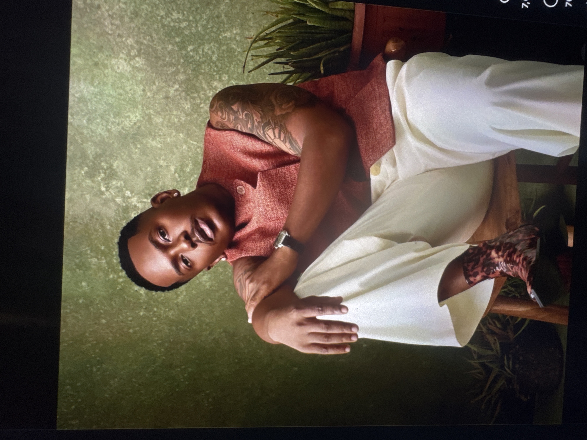 A man is seated on a wooden stool, his body angled towards the left of the frame. He is wearing a red vest over a white shirt, and loose-fitting cream-colored pants. His left arm is bent, with his hand resting on his leg, while his right arm is crossed over his chest. He has short, dark hair and is wearing earrings and a watch. His legs are crossed, and he is wearing patterned boots. Behind him, a textured green backdrop is visible, with potted plants to his right and left. The overall scene is a studio portrait. There is no indication in the image that this scene is located in Abuja, Nigeria.