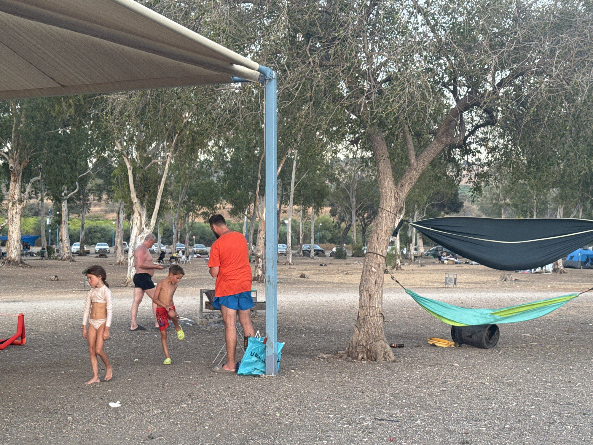A group of people are at a campsite near trees in the Golan Regional Council, Israel. In the foreground, a young girl in a light-colored swimsuit stands on a gravelly surface. To her right, a young boy wearing red shorts and yellow sandals is partially visible. An adult male in an orange t-shirt and blue shorts stands further back, near a barbecue grill. A large blue bag is on the ground next to a blue pole. Two hammocks are strung between trees in the background, one dark green and one turquoise. Cars are parked in the distance, and a tent is visible behind the hammocks. The ground is covered in small stones and gravel.