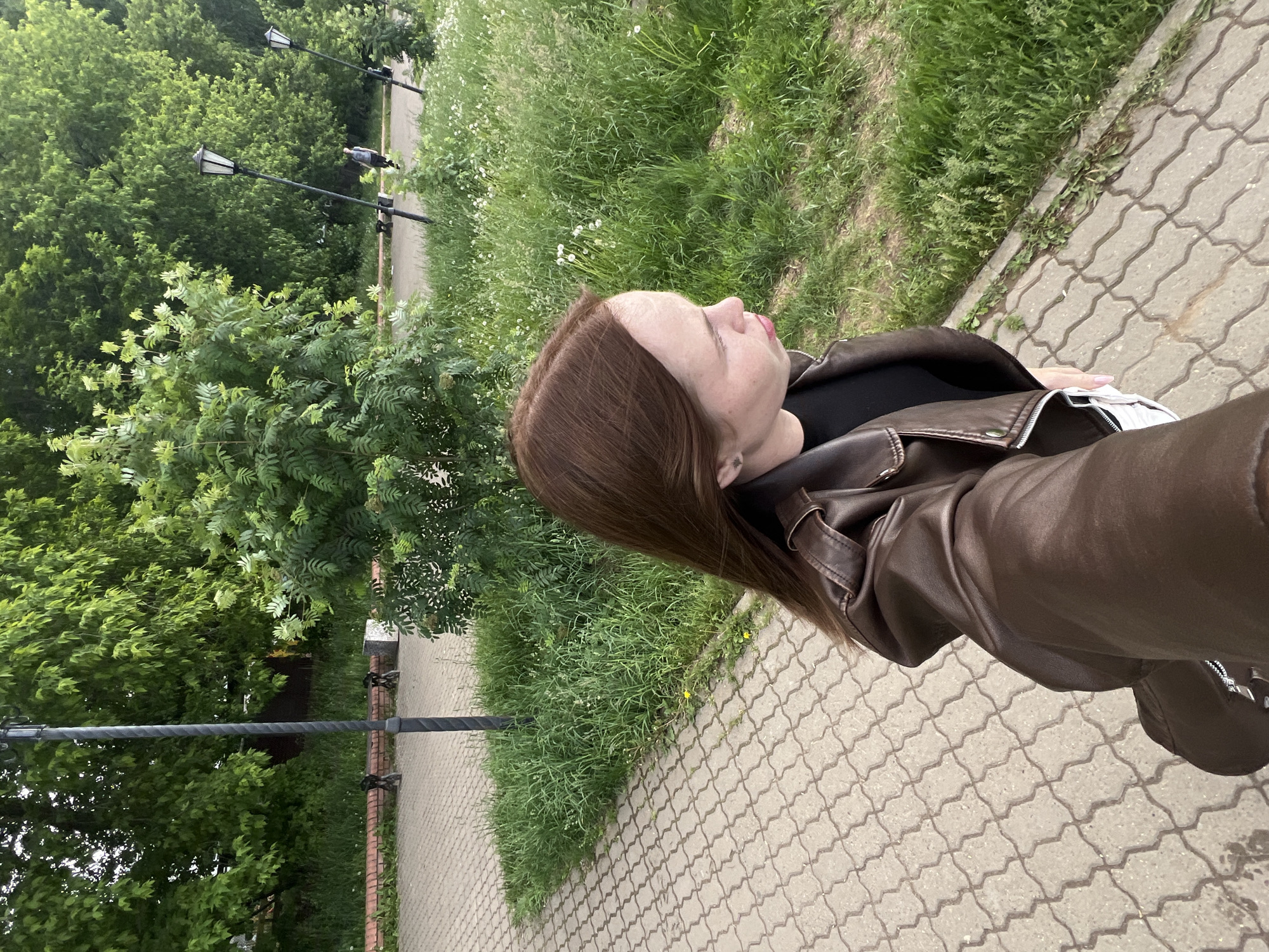 An outdoor, side-angle self-portrait features a young woman amidst a verdant park or pedestrian area in Chaikovsky, Russia. The woman, with long, straight brown hair, is wearing a dark brown leather jacket over a black top. Her profile is visible, looking slightly forward and up, with a neutral expression and lips slightly parted. She appears to be holding the camera in her right hand, capturing herself and the environment.
The setting consists of a paved path made of interlocking gray concrete pavers, which runs from the foreground into the background. Flanking the path is lush green grass, dotted with small white and yellow wildflowers. Beyond the grass, dense green trees and bushes create a rich, natural backdrop, indicating a vibrant summer or late spring environment. Two black, classic-style streetlights with decorative tops are visible along the path in the mid-ground, suggesting a public amenity.
The lighting is soft and diffused, consistent with an overcast day, or possibly early morning or late afternoon, as there are no harsh shadows. The overall mood of the image is calm and natural. No discernible text is visible anywhere in the image.