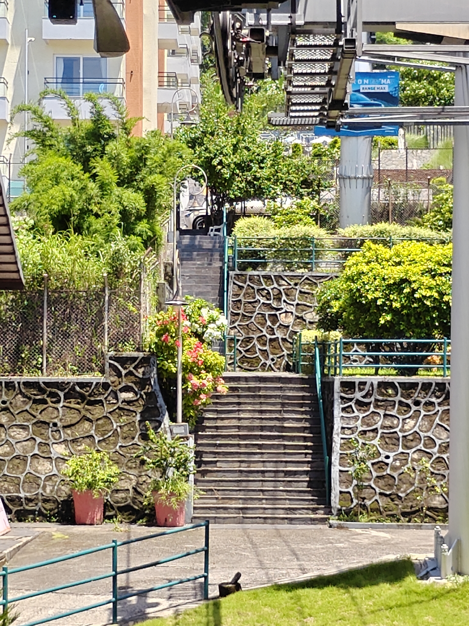 The image depicts an outdoor scene in Pokhara, Nepal, focusing on a section of the city's cable car system. In the foreground, a metal railing borders a grassy area. Behind this, a paved pathway leads to a wide, stone staircase that ascends a hillside. The stairs are flanked by decorative stone walls with a geometric pattern. Lush green vegetation, including flowering bushes with pink and white blossoms, grows alongside the stairs and on terraces. In the background, the cable car infrastructure is visible, with its track and support structures. Beyond the vegetation and cable car, parts of a multi-story building with balconies are visible. The scene is bathed in bright sunlight, suggesting a clear day. The image is a still, candid shot, capturing the architectural elements and natural surroundings of the cable car station.