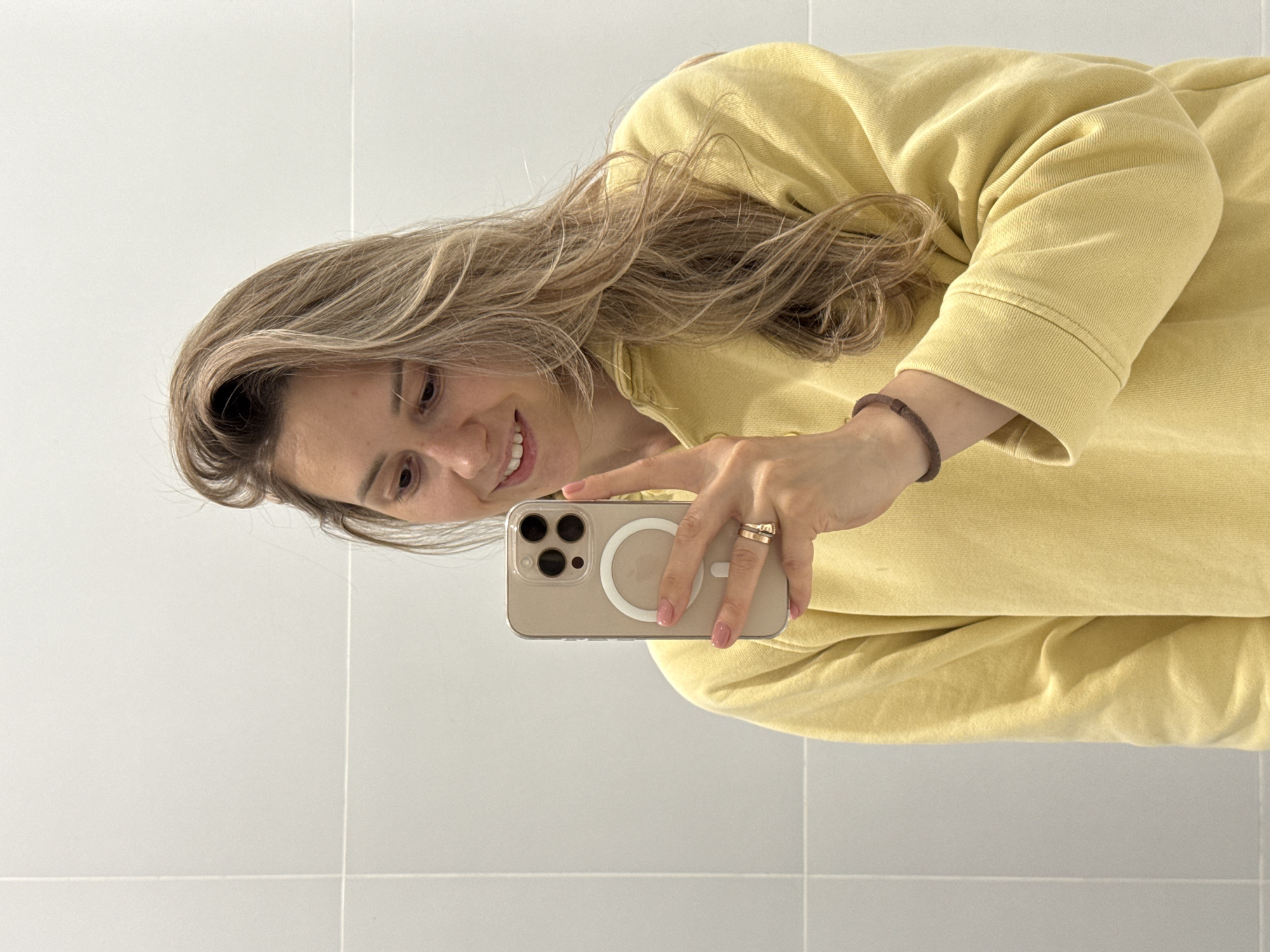 This mirror selfie, currently rotated 90 degrees counter-clockwise, features a smiling woman with long, wavy light brown hair. She is wearing a mustard yellow long-sleeved top, possibly a sweatshirt. On her left wrist, she has a dark brown beaded or woven bracelet, and on her right ring finger, she wears two rings. Her fingernails are painted a soft pink.
The woman is holding a light gold or champagne-colored smartphone, likely an iPhone, encased in a clear protective cover. A distinctive white circular MagSafe ring is prominent on the back of the phone, partially obscuring the Apple logo. Her gaze is directed towards the phone screen, indicating she is actively taking the picture.
The background consists of light-colored, possibly white, tiled walls with visible grout lines, suggesting an indoor setting such as a bathroom or a modern room. The lighting appears bright and even. The overall impression is one of casualness and a pleasant mood, conveyed by her gentle smile. The provided location context for this media is Новая Казмаска, Russia. No specific time of day or weather is discernible.