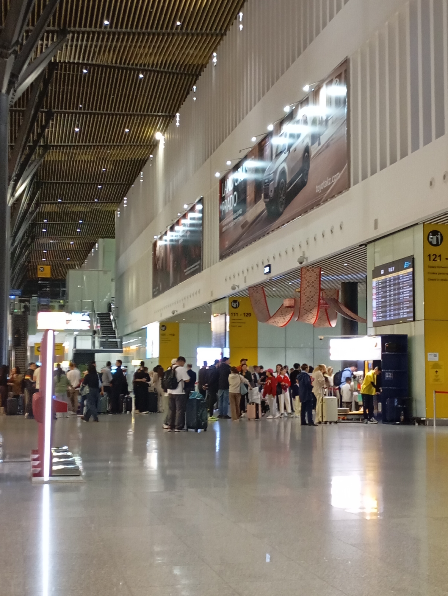The image depicts the interior of a modern airport terminal, likely in Astana, Kazakhstan. The scene is a busy concourse with numerous travelers queuing at a check-in area or information desk.
In the foreground, the polished, reflective floor shows streaks of light and shadows. Several people are standing in line, some with luggage. A prominent red and white information pillar with a digital display is to the left. Further into the terminal, a group of passengers, dressed in casual attire, are clustered around a check-in counter. A person wearing a yellow vest, possibly an airport employee, is attending to passengers.
The architecture is characterized by a high ceiling with a distinctive wooden slatted design and linear lighting fixtures. Large advertisements are mounted on the walls, one featuring a silver SUV, identified as a Toyota Land Cruiser Prado, with the website toyotakz.com. Yellow signage with flight information or gate numbers is visible, indicating areas for passengers. Escalators and staircases are visible in the background, suggesting multiple levels within the terminal. The overall atmosphere is one of transit and activity, typical of an international airport.