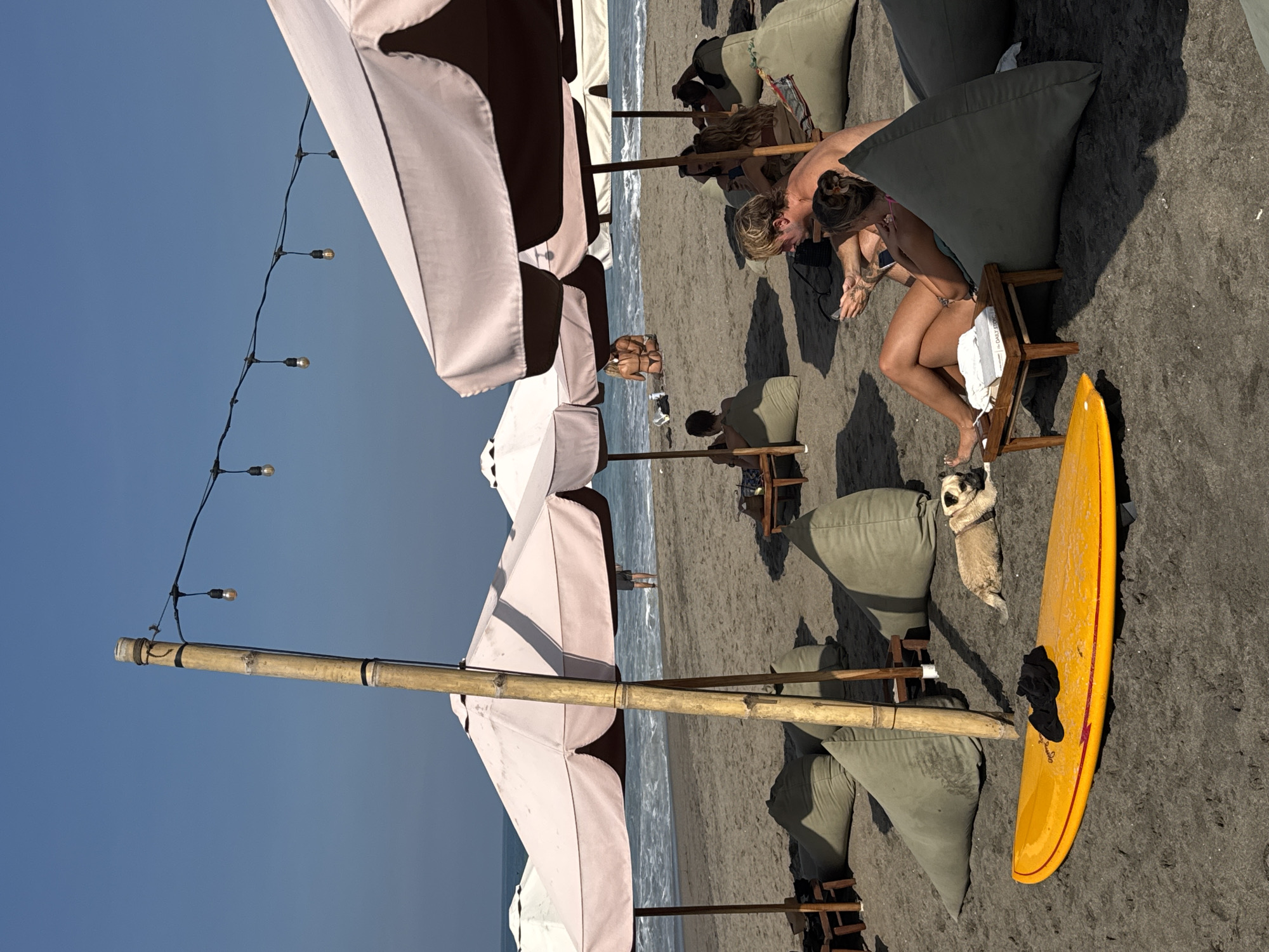 The image shows a beach scene in North Kuta, Indonesia. The location is set up with white umbrellas, wooden poles, and string lights overhead. Several people are seated on beanbag chairs at the beach. A woman in a bikini is sitting on a wooden chair, and a tan pug dog stands near her. A yellow surfboard is standing next to the chair. The beach is sandy and the ocean is visible.