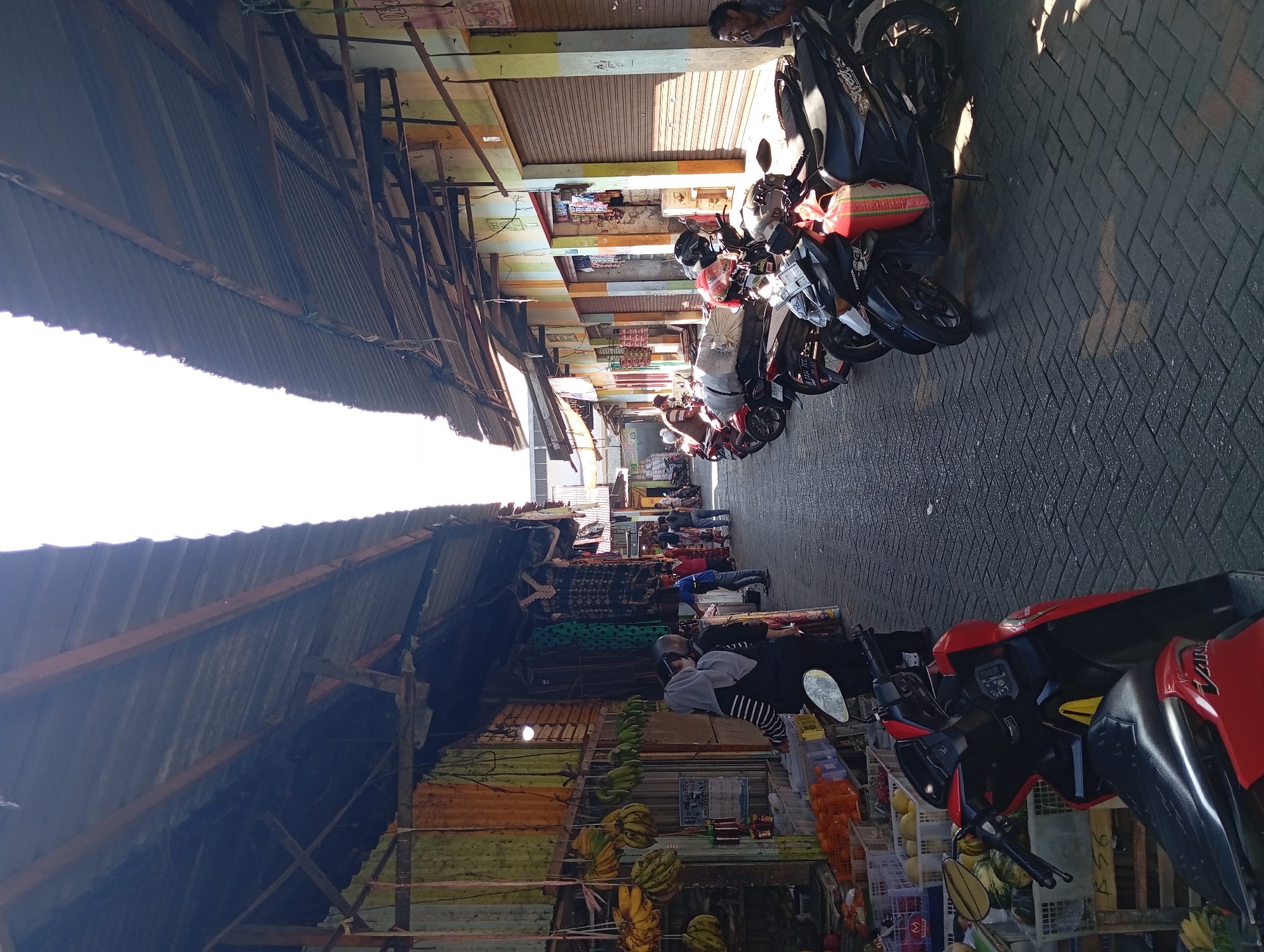 The image depicts a busy marketplace in the city of Kepulungan, Indonesia, on a bright, sunny day. The scene is characterized by a narrow pathway lined with market stalls on both sides, covered by corrugated metal roofs supported by wooden beams.
On the left side of the pathway, market stalls display an assortment of goods, including bunches of bananas hanging from the ceiling, various fruits and vegetables in crates, and clothing items like dresses and hijabs. Several people are browsing or tending to the stalls. A woman wearing a grey hijab and black and white striped shirt is seen from the back, with a scooter parked in front of her. Another woman in a grey hijab and black outfit is also visible.
On the right side of the pathway, a row of motorcycles is parked along the market stalls. These motorcycles appear to be modern scooters. The market stalls on this side have brightly painted facades in yellow and orange, with closed roller shutters indicating shops that may be open or closed. A man is seated on the ground near a motorcycle, appearing to observe the activity.
The ground of the pathway is paved with dark grey interlocking bricks. Sunlight streams through the gap between the two rows of market roofs, casting strong shadows on the pathway and the parked motorcycles. The overall atmosphere suggests a typical bustling Southeast Asian market.