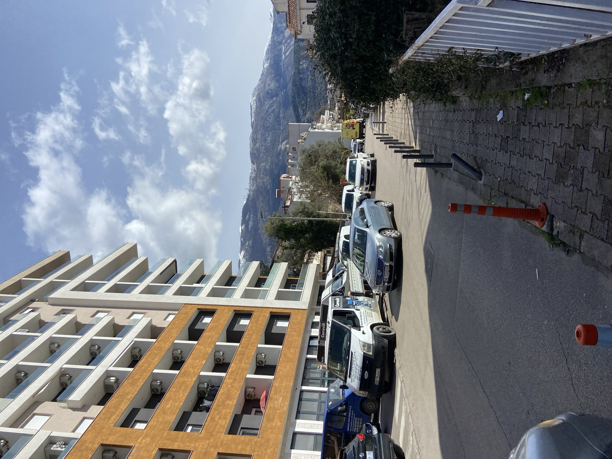 The image depicts a street scene in Budva, Montenegro, under a partly cloudy blue sky with bright sunlight.
In the foreground, an asphalt road occupies the center and left, showing visible cracks. A paved sidewalk made of grey interlocking concrete blocks runs along the right edge, bordered by a line of black, cylindrical bollards. An orange and white striped flexible traffic cone stands on the sidewalk in the immediate foreground, with a smaller orange bollard further back. A white metal fence and a dense green hedge are located to the right of the sidewalk.
On the left side of the road, several vehicles are parked or traveling. A dark blue van is partially visible on the far left. Adjacent to it, a light blue commercial van displays "ARMIRA MONTANA D.O.O." and a Budva license plate "BD-408-AX". To its right, a white Ford Transit commercial van with "ARMIRA MONTANA D.O.O." and "Ceresit" branding bears a Budva license plate "BD-BJ443" and features white objects on its roof. A silver-blue Volkswagen Passat sedan with Montenegro license plate "NK-EA639" is positioned centrally on the road, indicating movement. Further down the street, a white SUV and a yellow delivery van are visible.
A prominent multi-story residential building dominates the left side of the frame. Its lower section features a brown/orange facade, while the upper floors are lighter in color. Balconies with glass railings are present on each floor, equipped with numerous external air conditioning units. On one of the lower brown-section balconies, a red patio umbrella is open, and clothes are visible drying. The ground floor of this building contains large windows, suggesting commercial or retail spaces.
In the midground and background, additional buildings of varying sizes and colors are visible, interspersed with green trees. The distant background is characterized by a range of prominent, snow-capped mountains under the partly cloudy sky. Shadows on the road and sidewalk indicate the sun is positioned to the right or front-right of the frame. No individuals are explicitly visible.
