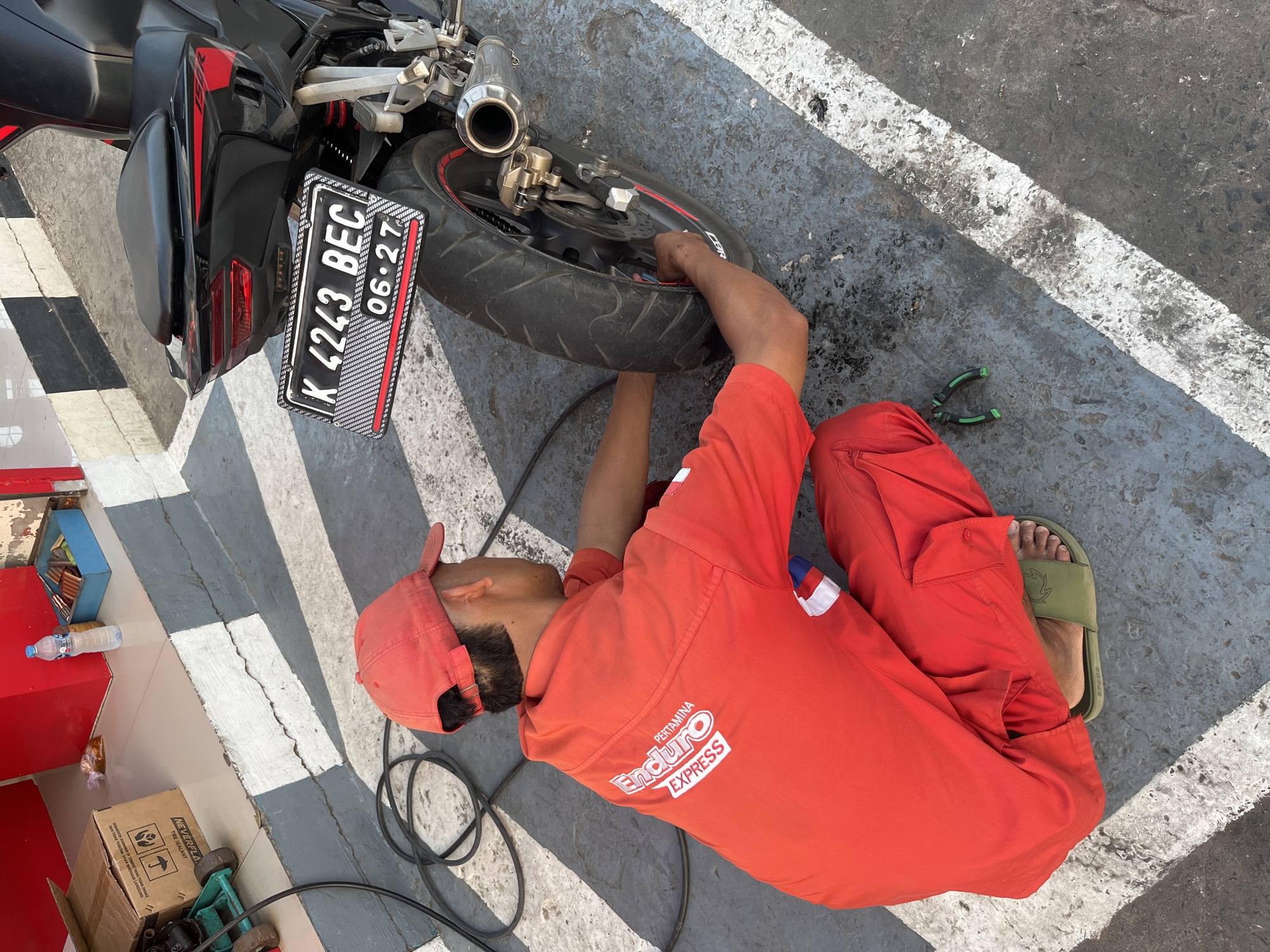A person in red work clothes and a red cap is crouched down, working on the rear tire of a black motorcycle. The motorcycle has a license plate that reads "K 4243 BEC 06-27." The person is using tools to work on the tire. The scene appears to be an outdoor maintenance area, with a gray concrete ground marked with white lines. In the background, there are red structures and a cardboard box. The location is identified as Pecangaan, Indonesia.