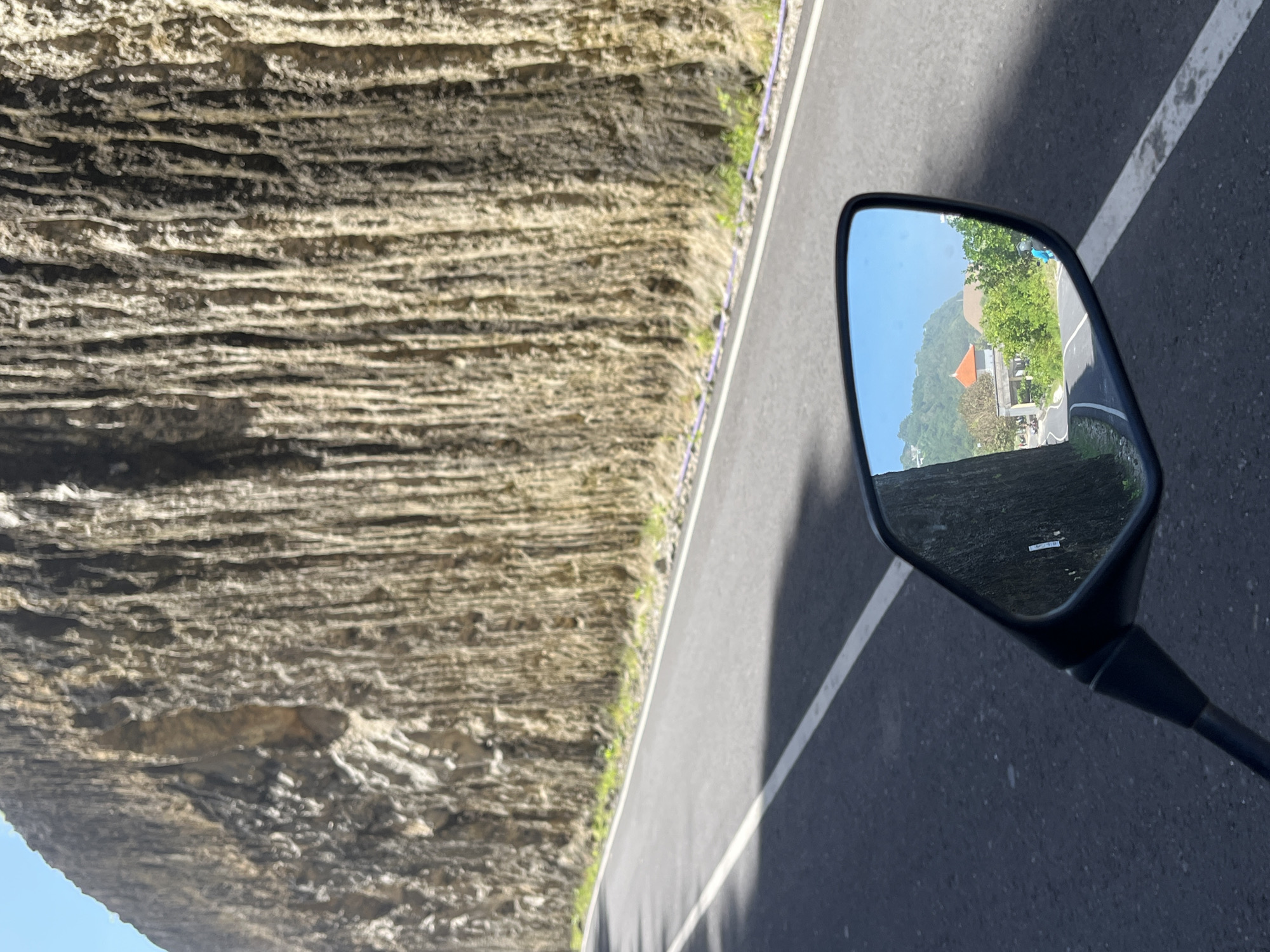 A motorcycle's side mirror reflects a building with an orange roof and green foliage in Kuta Selatan, Indonesia. The view outside the mirror shows a gray asphalt road with a white line and a tall, layered rock wall on the left.
