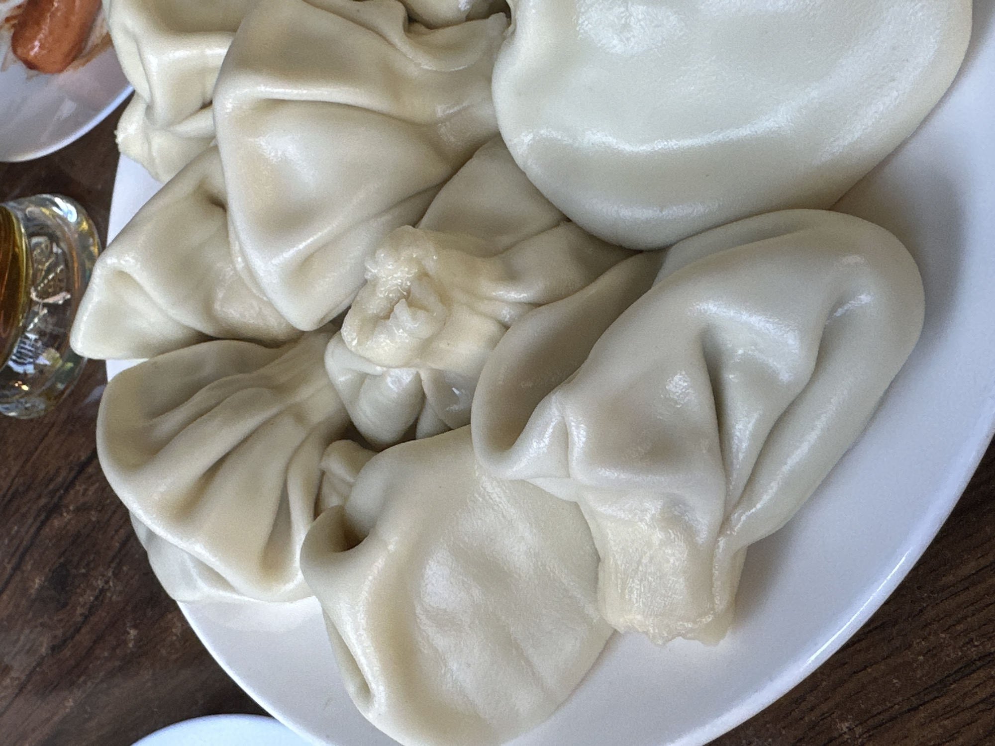 A close-up, high-angle shot captures a white plate piled high with numerous Khinkali, traditional Georgian dumplings. The dumplings are plump, with a pale, off-white color and a smooth, slightly glossy surface, suggesting they are freshly cooked and warm. Many display the characteristic pleated folds at their tops where the dough is gathered. This dish is commonly found in places like Tbilisi, Georgia, reflecting its culinary heritage.
In the upper left corner, a portion of another white plate is visible, holding a reddish-brown food item, possibly a sausage or another meat dish. To the left, a clear glass with a golden-hued liquid, possibly a beverage, sits on the table. The dishes are placed on a dark wooden table with visible grain, indicating an indoor dining setting. The image focuses on the culinary presentation, highlighting the texture and quantity of the Khinkali. No people or text are visible in the frame.