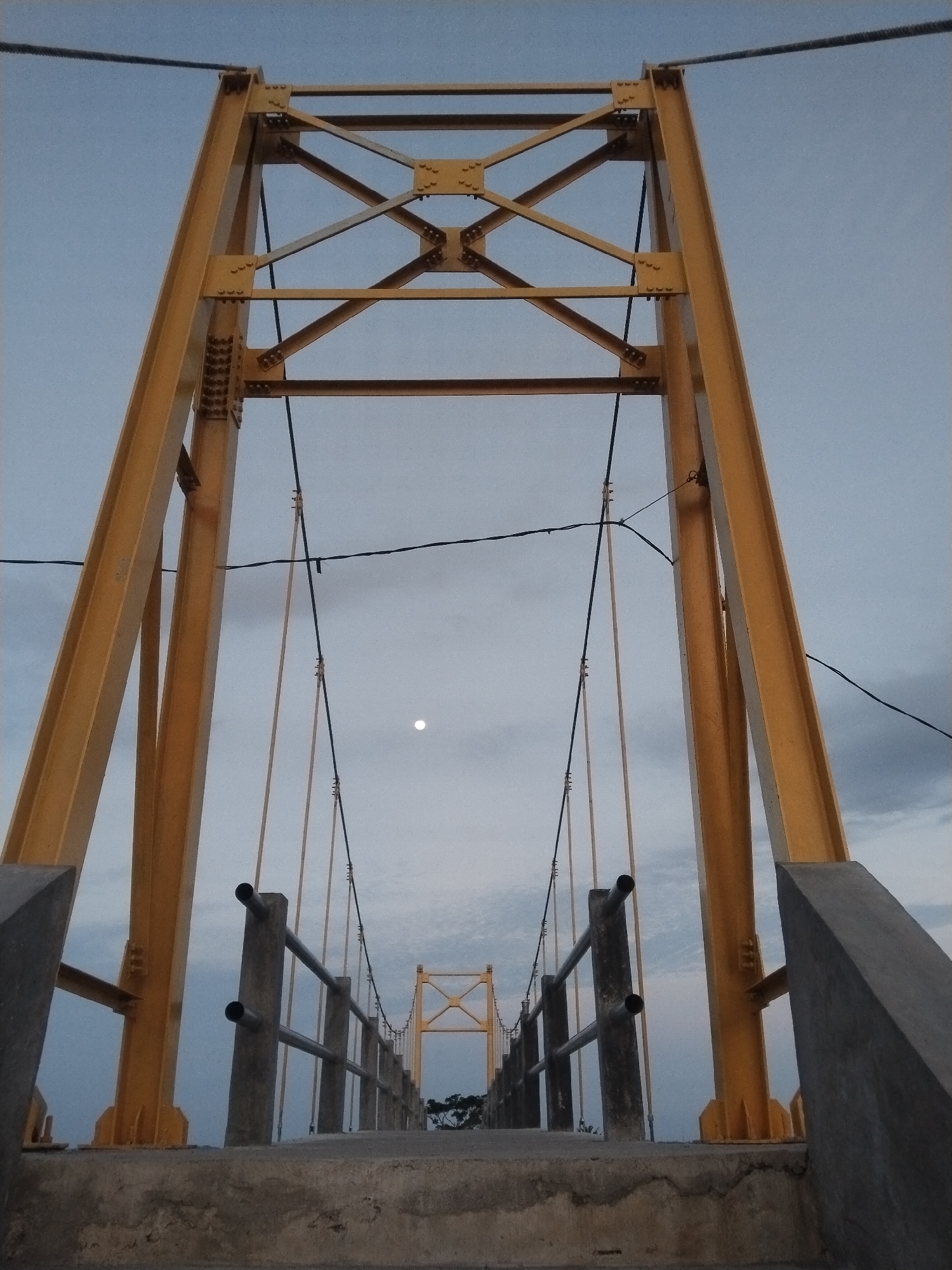 The image depicts a suspension bridge in Lhokseumawe, Indonesia. The bridge is constructed with yellow metal support towers and suspension cables. The deck is concrete with metal railing along the sides. The sky is overcast with a single white dot in the center, possibly the moon. The photograph is taken from the bridge deck, looking towards the opposite tower.
