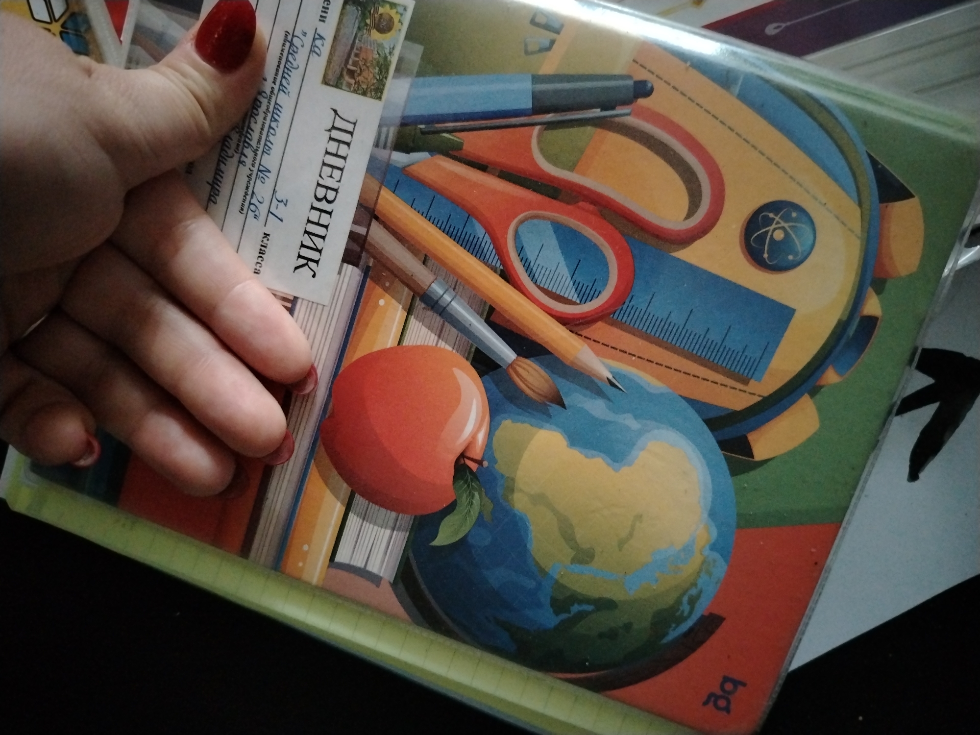 A person's left hand, with red nail polish, is resting on a school diary. The diary cover features a depiction of school supplies such as a backpack, scissors, ruler, pen, pencil, and a paintbrush, along with an apple and a globe. The diary also has a label that reads "ДНЕВНИК", which translates to "Diary" in Russian. The label also indicates "3-1 класса" (Grade 3-1), "Средней школы № 26" (Middle School No. 26), and then the location of "Ярославля" (Yaroslavl). The edge of other paper materials and black background are visible around the diary.