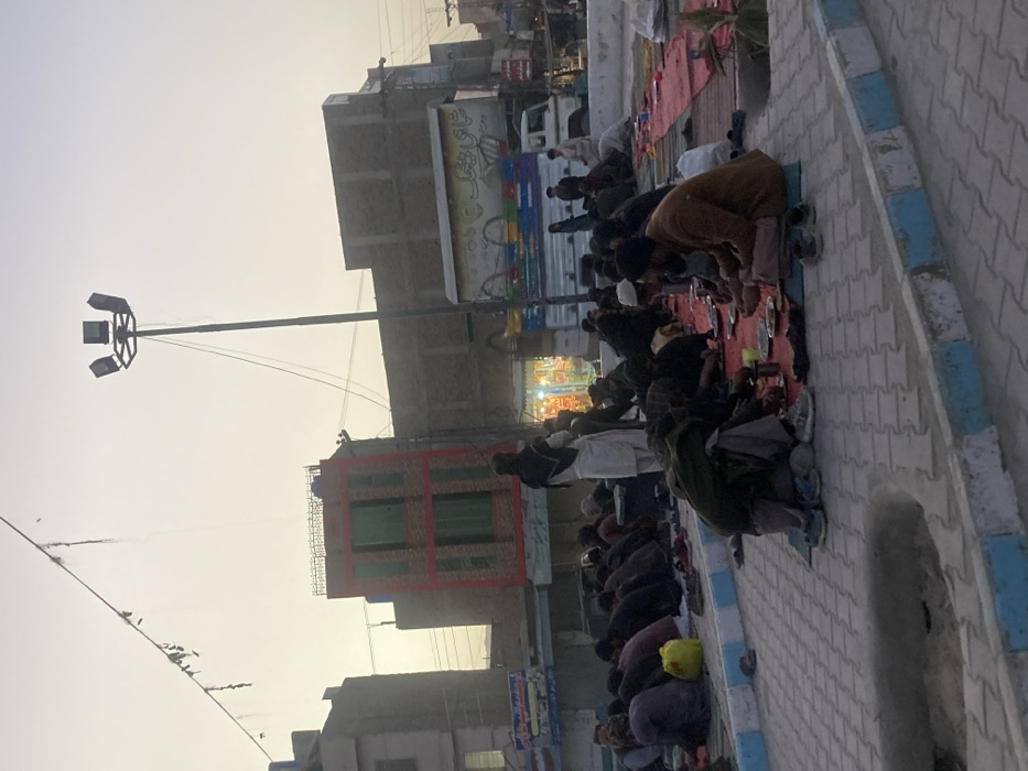 The image depicts an outdoor scene at dusk in Quetta City Tehsil, Pakistan. Approximately 30-40 adult males, dressed in traditional attire, are seated on red mats covering a paved area with interlocking pavers. Many are engaged in eating from individual plates/bowls. Footwear, including sandals and shoes, is removed and placed adjacent to the seated individuals. A yellow plastic jerrycan rests near the left edge of the seated group. A man in white traditional attire and a dark head covering stands to the left of center, observing.
The background features an urban street with multi-story buildings, including a prominent three-story structure with red brick facing and green-shuttered windows, and other buildings with concrete facades. A tall street light pole with four floodlight fixtures stands on the right. A white utility truck with colorful side advertising (including "شاہی کینک" in Urdu script) is positioned on the street behind the gathering. Several storefronts with illuminated interiors are visible. Overhead utility lines traverse the pale, dusky sky, beneath which distant hills are faintly visible. The foreground includes a light blue and white painted curb.