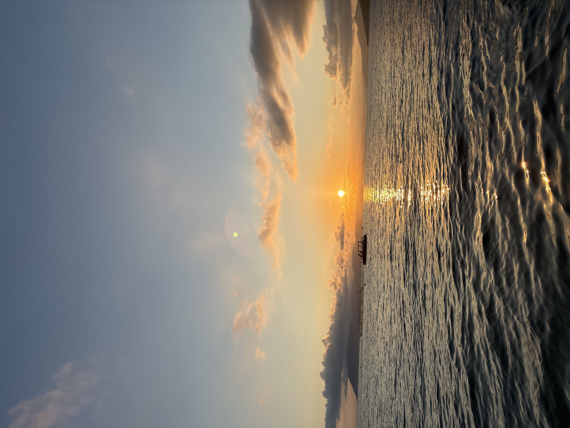The image captures a sunset over the sea, likely in Novorossiysk, Russia, as indicated by the context. The sun, a bright orange orb, is partially obscured by clouds on the left side of the frame, casting a shimmering golden reflection on the rippling water. The sky transitions from a pale blue at the top to warmer hues of orange and yellow near the horizon. Wispy pink and orange clouds are scattered across the sky.
In the distance, a dark silhouette of a boat is visible, moving across the water. Further in the background, landmasses with gentle hills are outlined against the sunset. A few other smaller boats or structures can be seen on the water in the far distance. The foreground is dominated by the dark, textured surface of the sea, with small waves creating a pattern of light and shadow. The overall scene evokes a sense of calm and natural beauty.