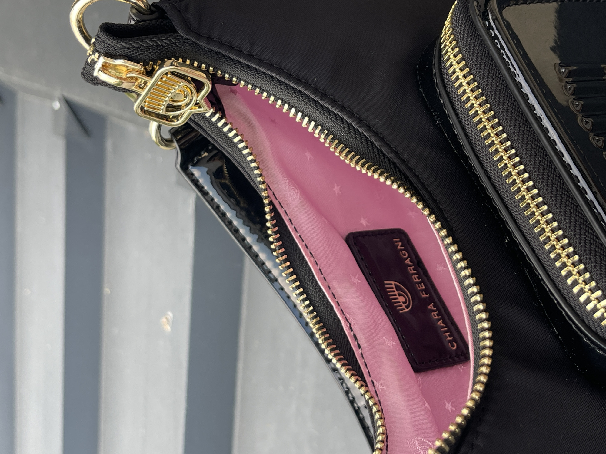 A close-up, high-angle shot focuses on the interior and exterior details of a black Chiara Ferragni handbag with a pink satin lining. The bag features a prominent gold zipper with a decorative gold charm shaped like the brand's iconic eye logo. A dark rectangular tag with the same eye logo and "CHIARA FERRAGNI" embossed in gold is attached to the interior lining. The lining itself has a subtle pattern of small pink stars. A portion of a second zippered compartment, also with gold hardware and a shiny black patent leather front, is visible at the bottom of the frame. The background consists of blurred gray horizontal lines, resembling steps or seating. The image does not contain any people or provide any information about the city of Karlovo, Bulgaria.