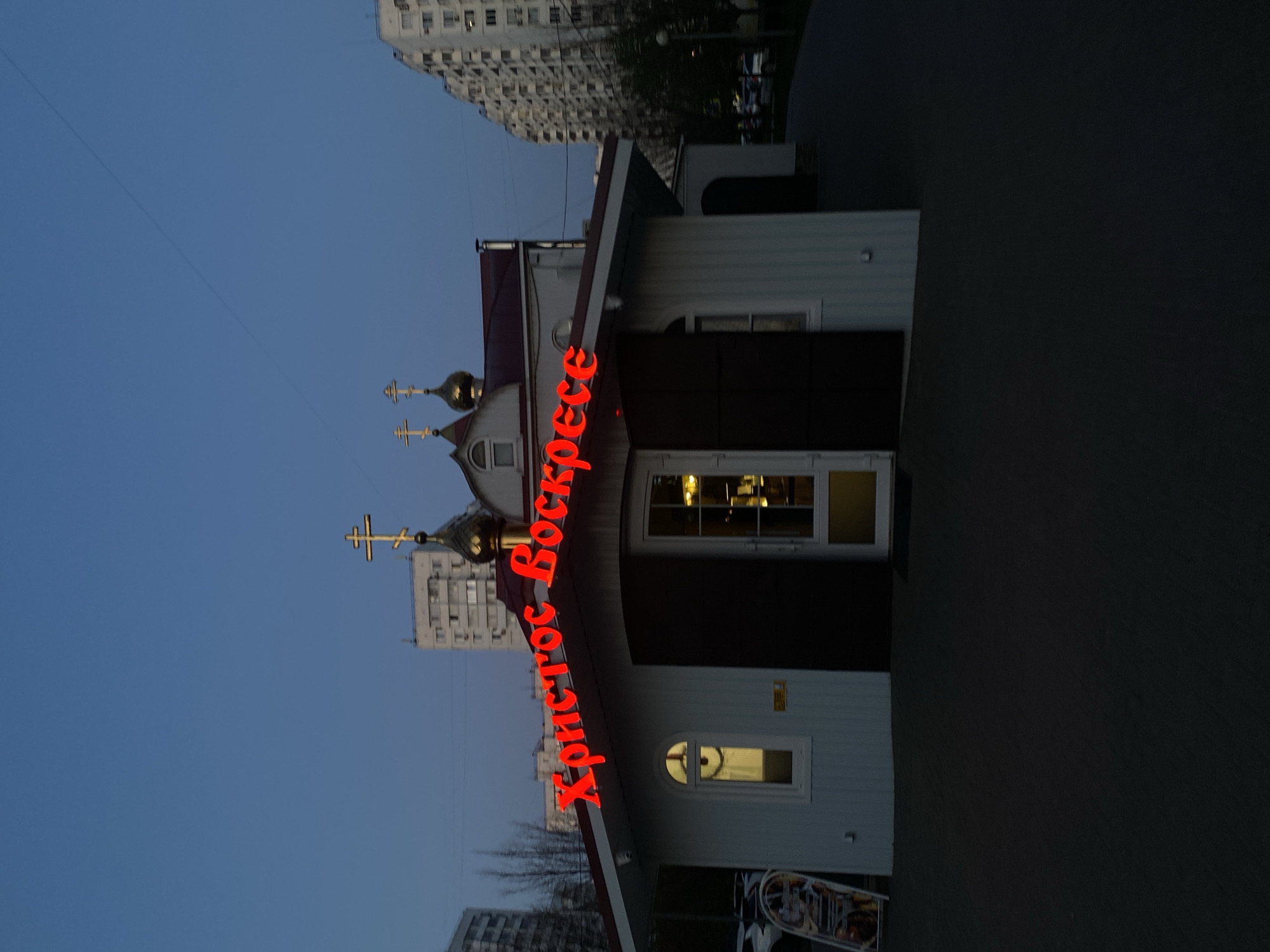 Captured from a low-angle perspective, the image features a church building against a backdrop of a clear, blue evening sky and a glimpse of urban high-rise buildings. The church is adorned with traditional golden domes and crosses, and lit up by a red neon sign reading "Христос Воскресе". The environment is a mix of urban architecture and religious elements, indicating a city setting, but the specific location is not identifiable from the provided information.