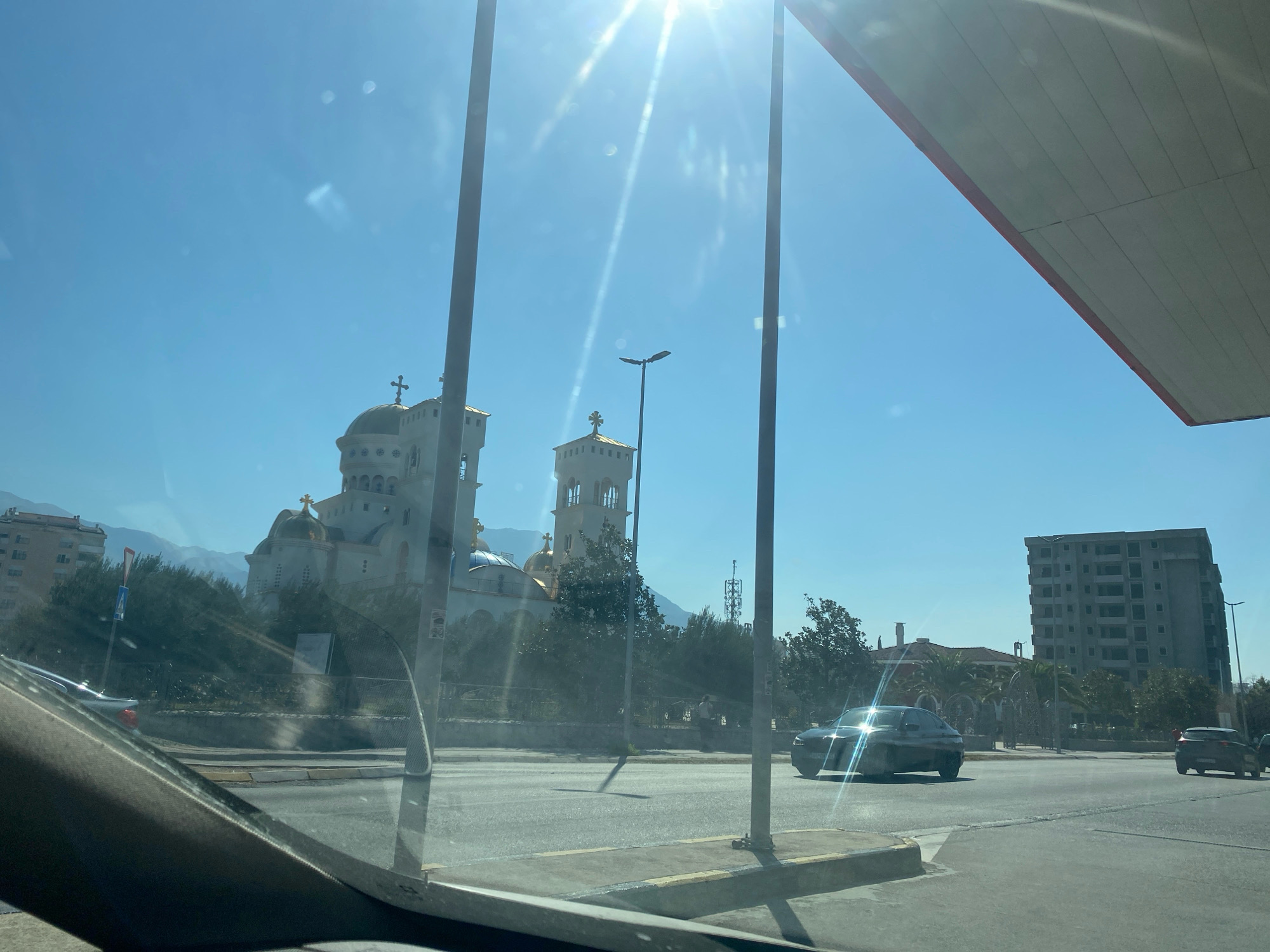 In the city of Bar, Montenegro, on a clear sunny day, the view through a vehicle windshield reveals an Orthodox church with golden domes and crosses on the left, a large unfinished building on the right, and palm trees scattered throughout. A road with several cars is in the foreground. The sun is bright, causing glare and lens flares.