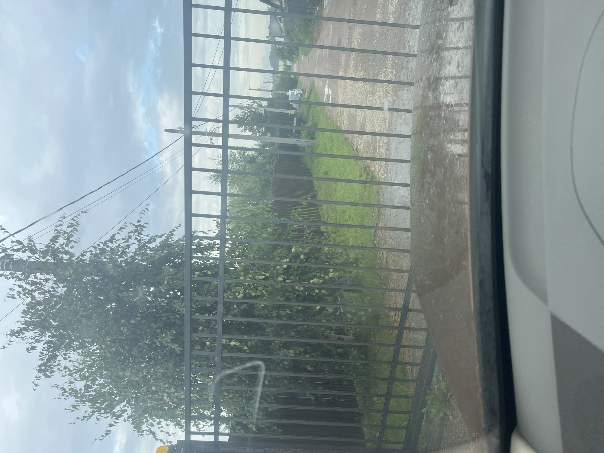 The image shows a view through a metal fence into a rural or suburban area. A dirt road leads away from the viewer, with houses and trees visible on either side. A white car is parked on the right side of the road. The sky is overcast, and it appears to have recently rained, as there are puddles on the road. The foreground of the image is dominated by the car's dashboard and windshield, with the fence in the middle ground and the scenery beyond. There are no people visible in the image. The location is described as Vladimirovka, Russia.