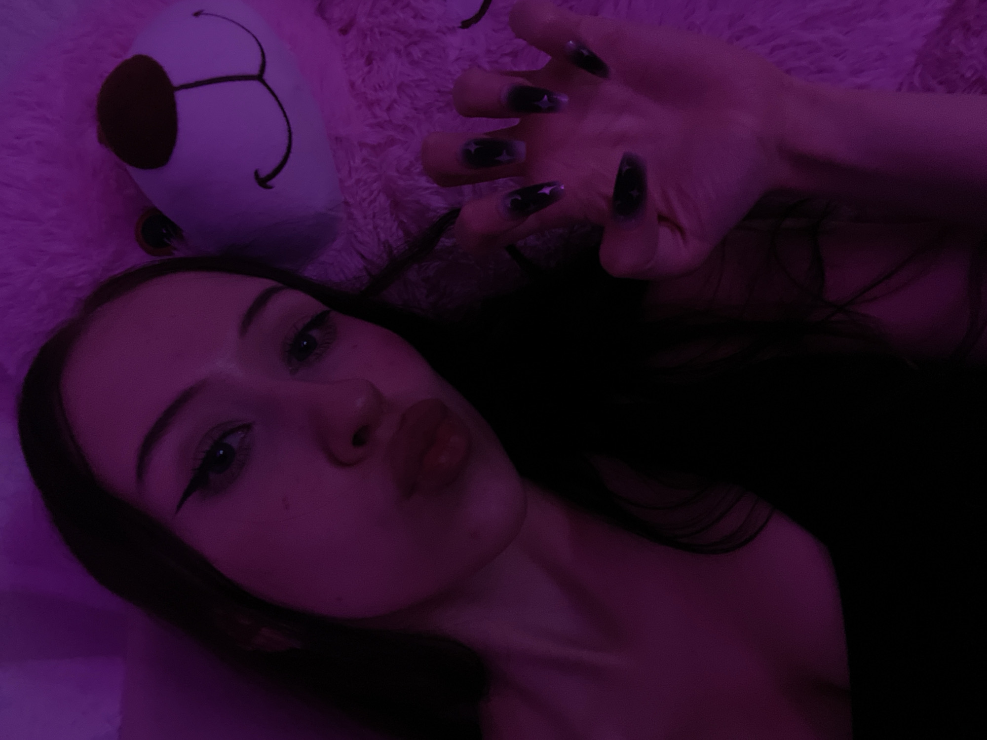 The image shows a close-up of a young woman lying down. She has long, dark hair and is wearing black eyeliner. Her lips are pursed. Her left hand is raised, showing long, dark nails with star designs. She is lying on a fluffy pink surface, and next to her head is the face of a white teddy bear with a brown nose and a smiling mouth. The scene is lit with purple light. The image is not provided with a location in Votkinsk, Russia.