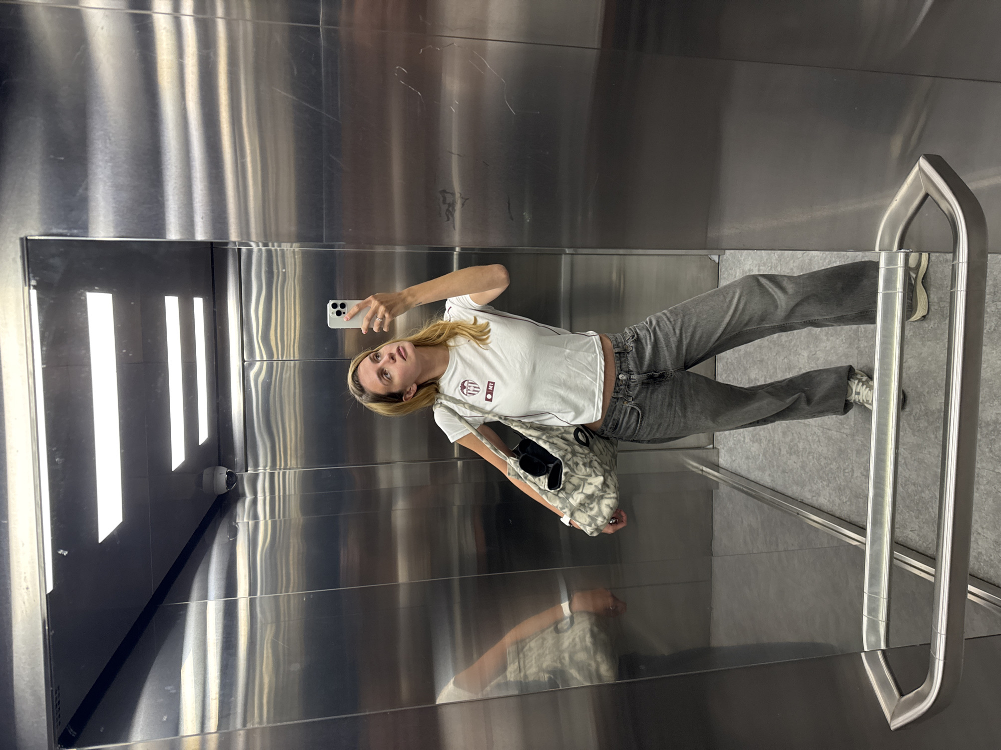 A young person, appearing to be female with blonde hair, is captured in a selfie taken in what appears to be an elevator. The image is presented as if rotated 90 degrees counter-clockwise, showing the subject standing upright against a highly reflective, brushed metal wall.
The person is wearing a white short-sleeved t-shirt with a small, circular, reddish-brown and white logo or emblem on the left chest area, which is difficult to read but seems to contain letters like "BMT" or "GMT". She is also wearing light gray jeans and light-colored sneakers. A patterned shoulder bag hangs from her left shoulder, with black objects visible inside. She holds a white smartphone with multiple camera lenses in her right hand, using it to take the photograph, and a ring is visible on her right ring finger.
The setting is characterized by shiny, metallic walls that reflect the subject and vertical strip lighting. A silver, U-shaped handrail is present on the right side of the frame, and the floor is a light gray. In the upper left corner of the main image, a white security camera is mounted on the wall, pointing inwards. The reflective surfaces create a mirrored effect, showing partial reflections of the subject and the internal structure of the elevator. The activity is clearly a selfie being taken within an enclosed space, likely an elevator. The time of day is not discernible due to artificial indoor lighting.