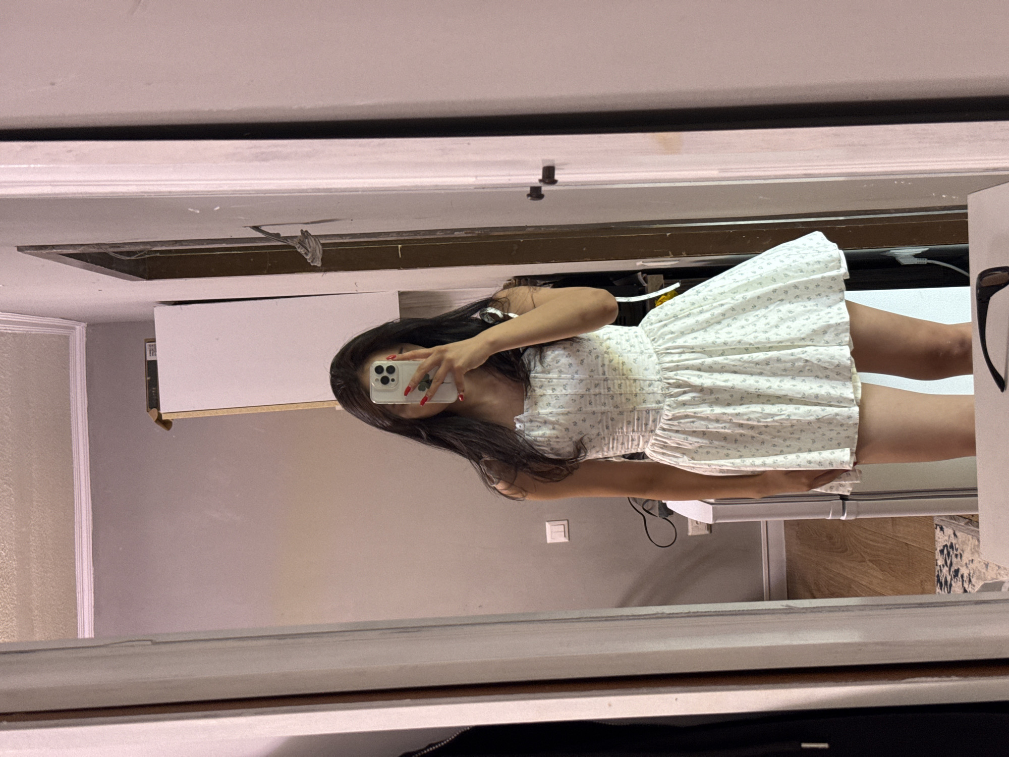 In Sary-Oy, Kyrgyzstan, a young woman with long, dark hair takes a mirror selfie in what appears to be a residential setting. She is wearing a white, floral-patterned, empire-waist dress with ruffled detailing. The dress has thin straps and a gathered skirt. Her nails are painted red. She holds a smartphone in her right hand, obscuring her face. The mirror reflects a room with pale walls, a light-colored cabinet or appliance, and visible electrical wiring. The floor is a mix of wood and a patterned rug. The overall impression is an informal snapshot in a domestic space.