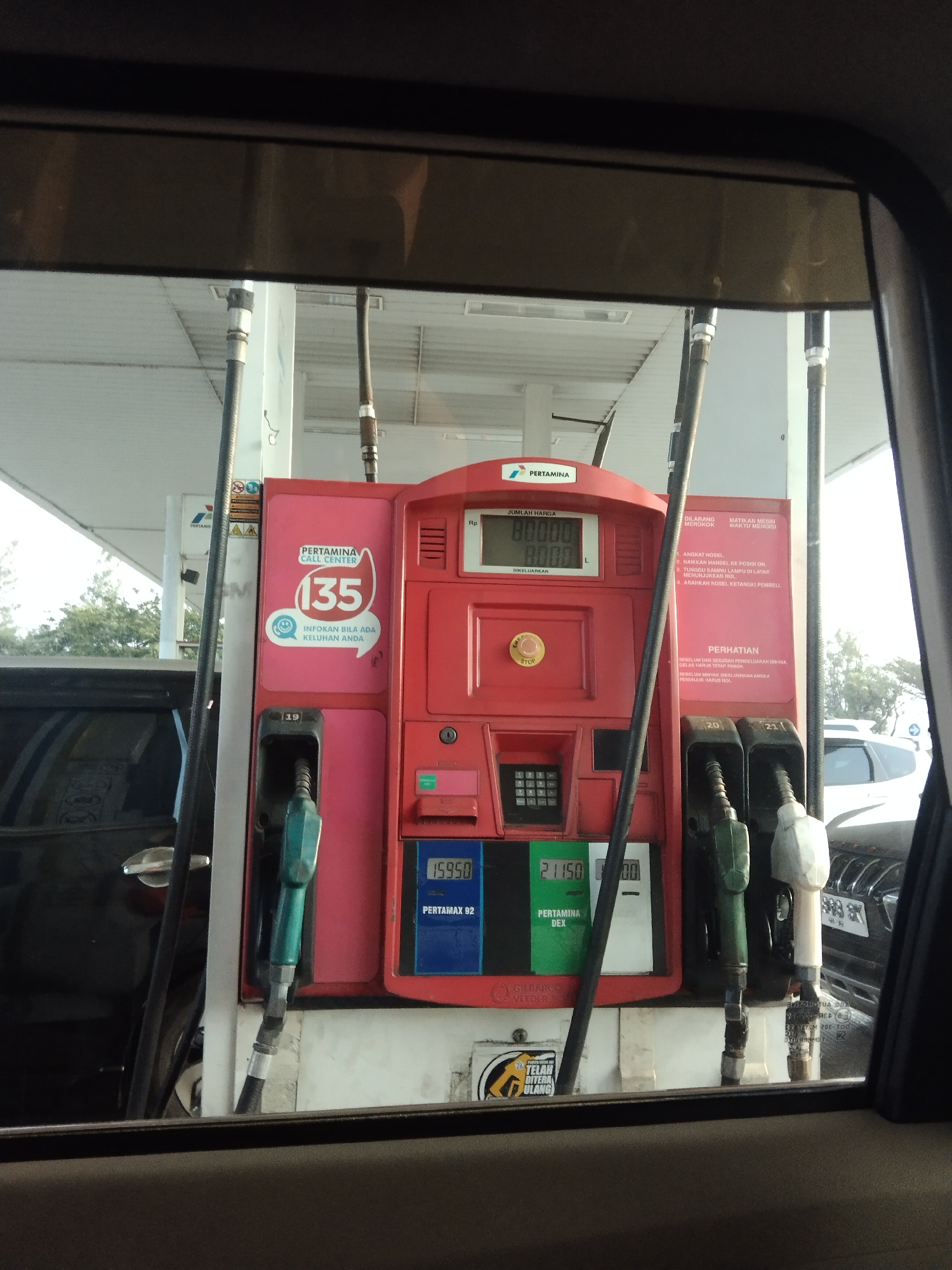 The image shows a red Pertamina gas pump at a gas station. A black car is parked next to the pump. The pump has several displays showing prices and quantities of fuel. There are also several nozzles for dispensing fuel. The scene is outdoors, and the sky is visible in the background. The image appears to be taken from inside another car, looking out through the window. The overall environment suggests a typical fuel stop in Indonesia.