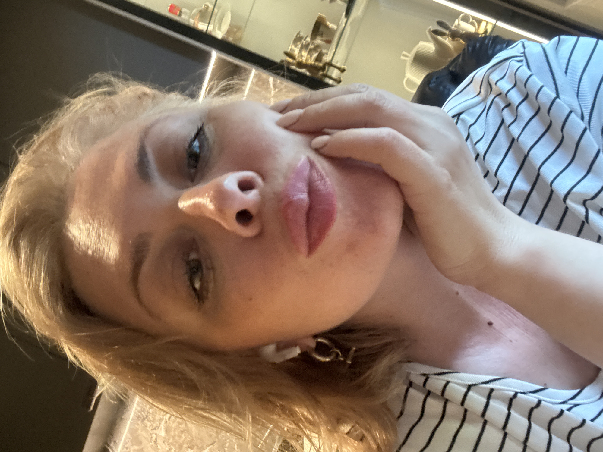 This is a close-up, selfie-style photograph of a woman with blonde hair, taken indoors. She is tilting her head and touching her face with her right hand, her fingers resting near her cheekbone and eye. Her lips are slightly parted and appear to have lip gloss on them. She is wearing a striped shirt with black and white lines. Her ear is adorned with a white earring with a gold chain detail. In the background, a shelf with decorative items, possibly in a cabinet or display case, is visible, illuminated by vertical lighting. The overall lighting suggests it could be daytime, but the interior setting makes it difficult to determine the exact time of day or weather. The image has a casual, personal feel, possibly capturing a moment of self-reflection or a quick photo. There is no visible text in the image.