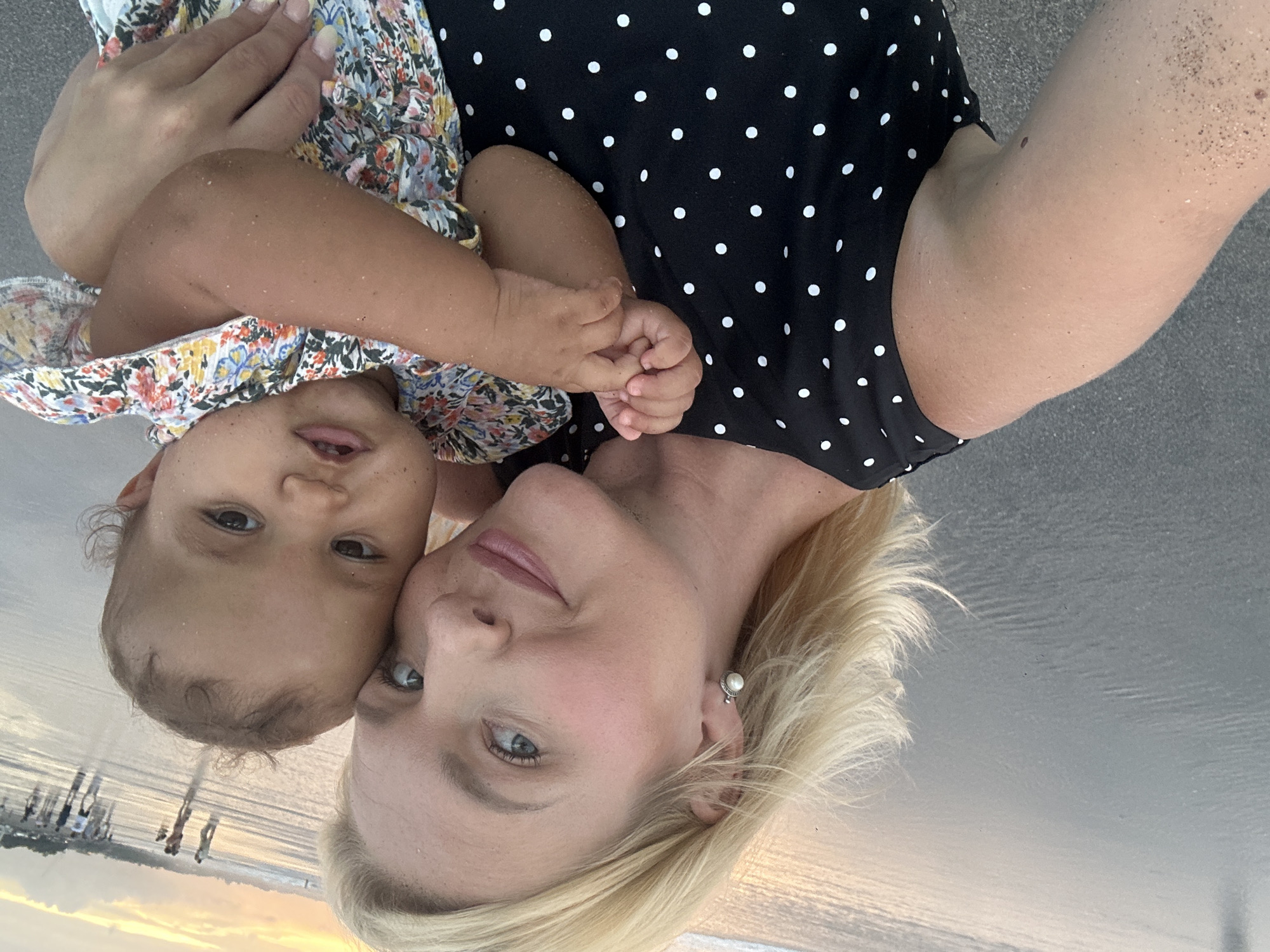 In the foreground, a fair-skinned woman with blonde hair is taking a selfie with a baby. She is wearing a black dress with white polka dots and a pearl earring in her left ear. The baby, with dark skin and curly hair, is being held by the woman and is wearing a floral-patterned shirt. The background shows a beach scene in North Kuta, Indonesia, with the ocean and other people in the distance. The sun is setting, casting a warm glow on the sky and the wet sand.