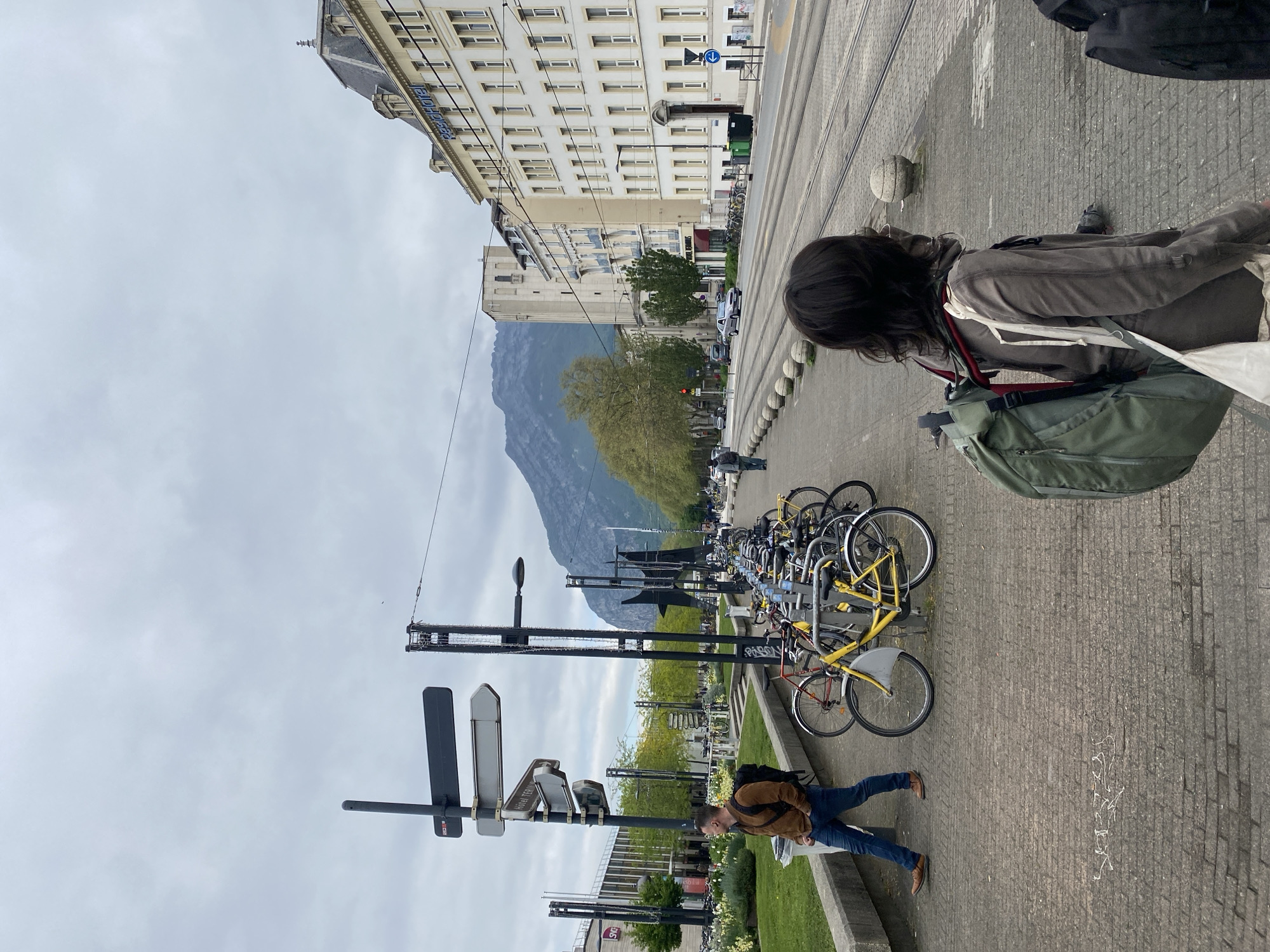 The image depicts an outdoor scene on a pedestrian walkway in Grenoble, France. The walkway is lined with tram tracks and stone ballards that run parallel to the sidewalk. A mountain looms in the background. The sky is overcast.
A row of parked bicycles are locked to a rack along the walkway. A woman stands on the walkway, with a large olive green backpack and dark hair. A man lies prone on the walkway. He wears blue jeans, brown shoes, and a brown jacket, appearing to be in a relaxed pose or possibly injured. There are signs indicating the name "Gare Terminus" and the "SNCF" logo. Buildings are present in the background with the name "RésidHotel" displayed at the top.