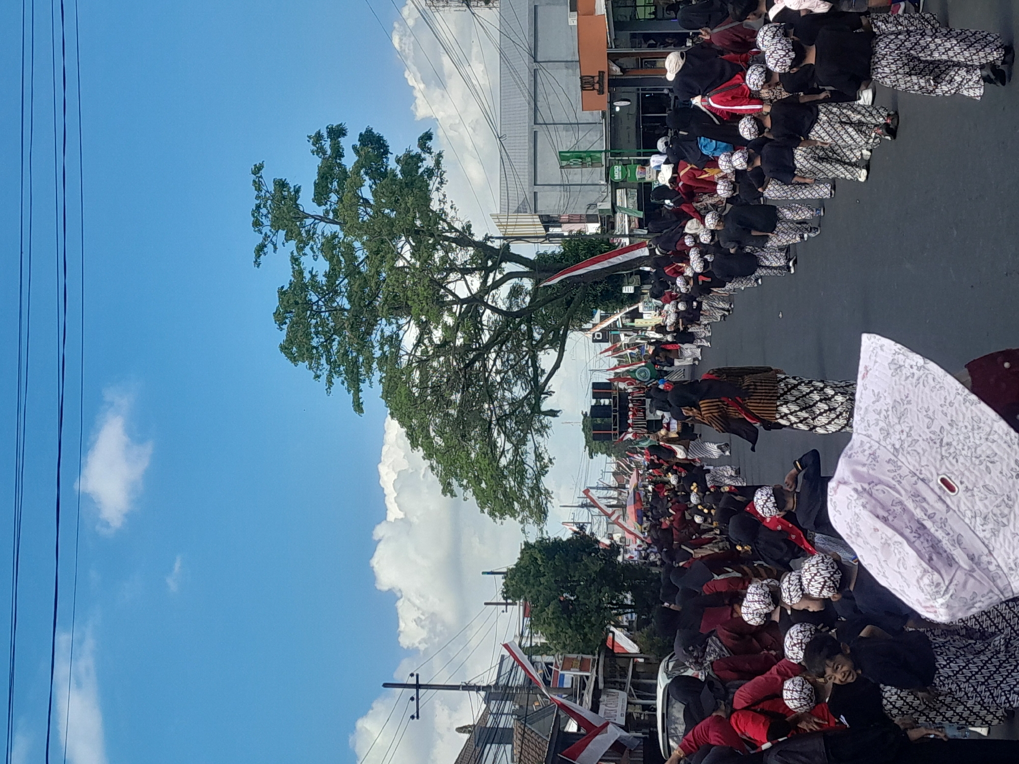 A wide shot shows a street procession in Genteng Kulon, Indonesia, under a bright blue sky with scattered white clouds. A large tree with lush green foliage dominates the center-right of the frame, its branches extending upwards. The street is lined with buildings and utility poles with power lines. Indonesian flags are prominently displayed on some buildings and poles. A crowd of people, mostly children dressed in traditional attire, are participating in the procession. Many of the children wear black tops and batik patterned pants and head coverings. Some adults, also in traditional dress, are visible amongst them, including some in red tops and batik skirts. A speaker system on a platform is visible in the background, suggesting a performance or ceremony. The overall scene depicts a community event or celebration.