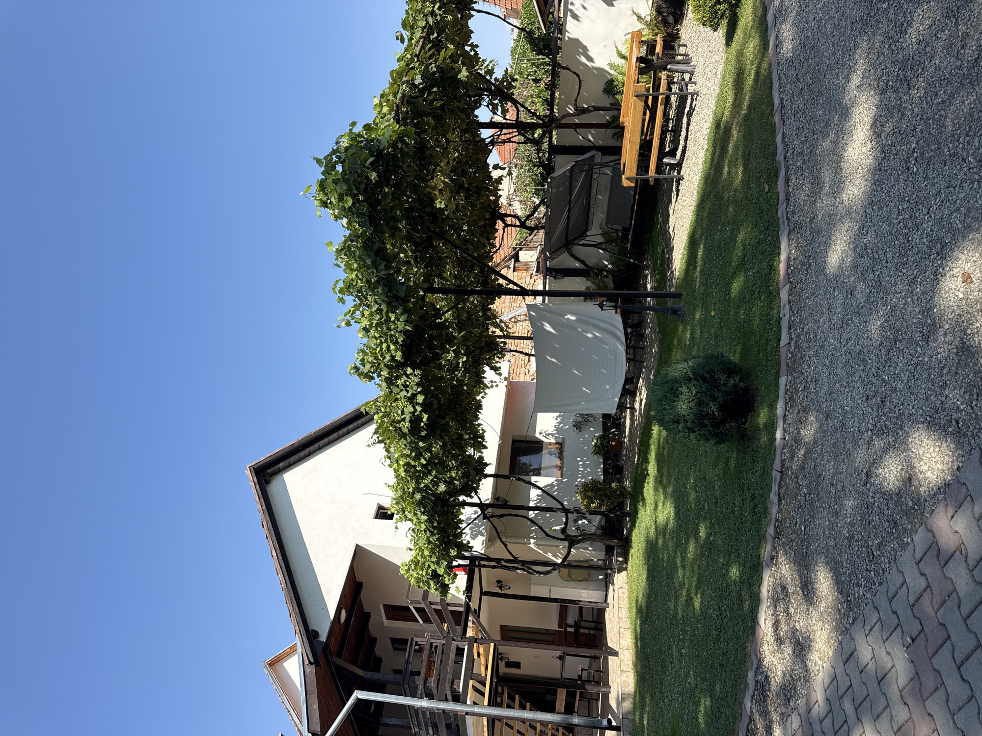 The image depicts a residential property on a sunny day in Cârțișoara, Romania. A two-story white house with a dark-tiled roof and wooden balconies is visible on the left side of the frame. An external metal staircase leads to the upper floor. The house is partially shaded by a vine-covered pergola that extends across the yard. In the foreground, a paved driveway with interlocking stones transitions into a gravel area. A well-maintained green lawn occupies the central part of the yard. Further back, a wooden picnic table with benches is situated on the gravel. Behind the table, a dark-colored swing set is present. A white sheet or cover is draped over a structure on the left side of the yard. The background shows other residential buildings with red-tiled roofs. The sky is clear and blue, indicating fair weather. There are no people visible in the image.