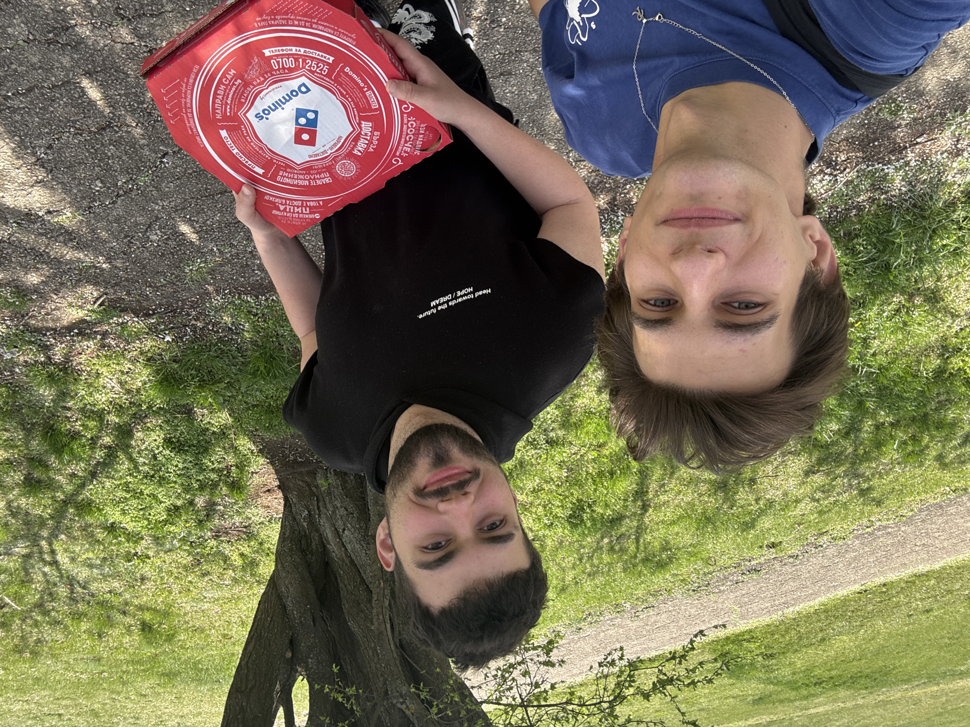 Two young men stand in a park in Sofia, Bulgaria. One holds a Domino's pizza box with Bulgarian text, including the phone number 0700 12525. The other wears a blue shirt with a necklace. The man holding the pizza box wears a black t-shirt with the text "Head towards the future. HOPE / DREAM". A tree is visible on the left, and a path and green grass are in the background. The image is taken from a slightly tilted angle.