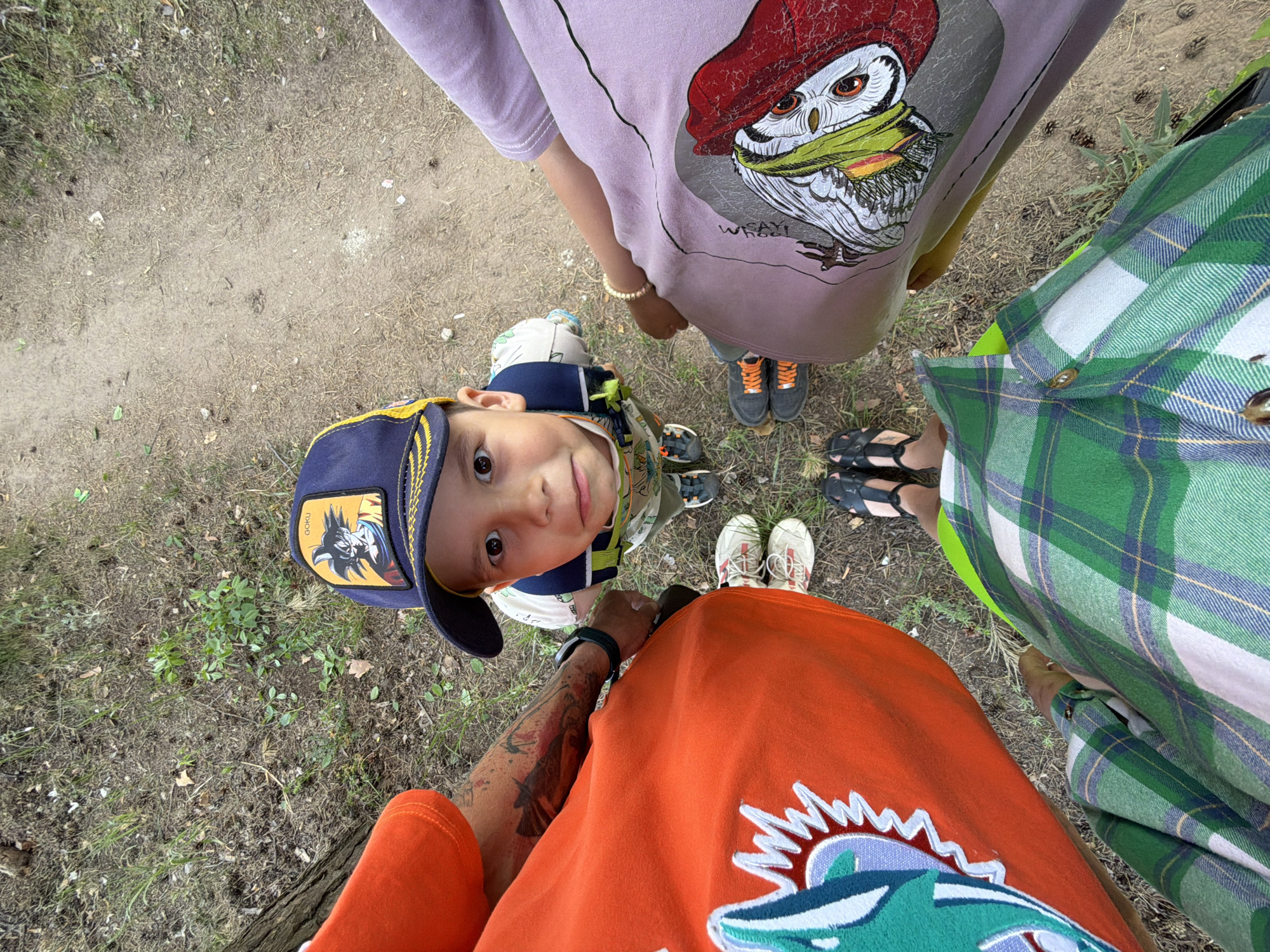 The image is a high-angle shot taken from the perspective of an adult, looking down on a child and two other people. The setting appears to be an outdoor area with bare earth and some scattered vegetation, suggesting a park or natural area, likely in Chaikovsky, Russia.
The child, wearing a blue baseball cap with a "Goku" character on it and a patterned shirt, is looking up at the camera with a slight smile. The adult directly beneath the camera is wearing an orange shirt with a blue and white design, and sneakers. A tattoo is visible on the adult’s arm.
To the side, another adult is wearing a lilac-colored shirt with a cartoon owl design, gray-black shoes and an accessory on the wrist. The third adult is wearing a green and blue plaid shirt and black sandals. The group seems to be standing in a circle. The overall impression is a candid snapshot of a group, possibly a family, spending time outdoors.