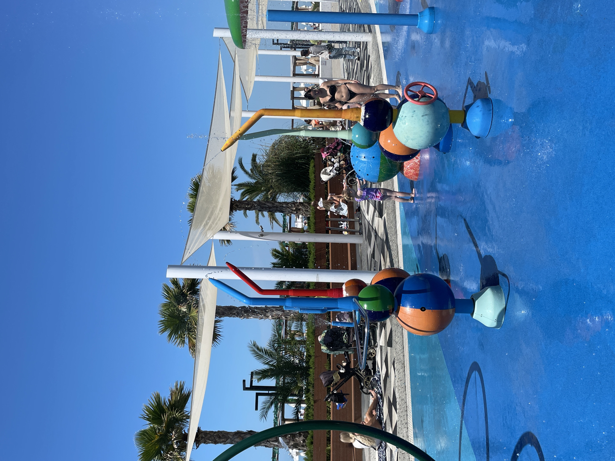 This image captures a lively outdoor splash pad area, likely part of a resort or recreational facility, under a clear, bright blue sky in Sochi, Russia. The time of day is daytime, with abundant sunshine indicating warm weather.
In the foreground and midground, colorful water play features are prominent, actively spraying water onto a vibrant blue, non-slip ground. These features include various sized spheres painted in blue, orange, and green, along with tall posts that emit water, and playful elements like a red steering wheel.
Several people are visible enjoying the amenities. An adult woman in a black bikini stands near the water features. Two young children are actively playing in the water; one child wears purple swim shorts. In the background, other individuals are scattered, some relaxing on lounge chairs, while others stroll on a patterned tile walkway. The setting is further defined by tall palm trees and large white triangular shade sails, providing a tropical ambiance and relief from the sun. Strollers are also present, suggesting the area is popular with families.
The overall scene conveys a cheerful and leisurely atmosphere, indicative of summer recreation and enjoyment. No visible text is present in the image.
