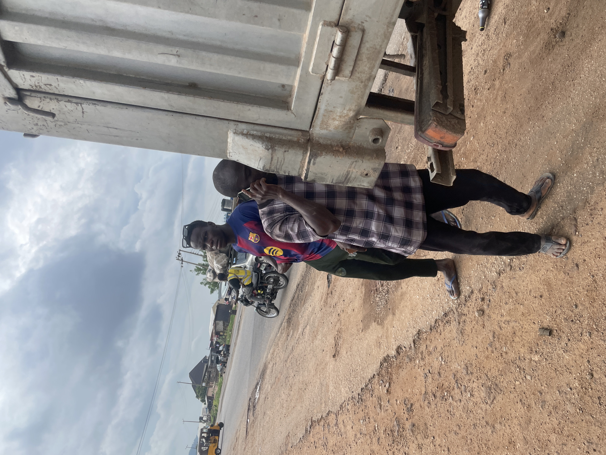 The image shows two men standing near a large, light-colored truck or trailer. The man in the foreground is wearing a plaid shirt over a blue and red jersey, black pants, and flip-flops. He is holding a phone to his ear. The second man, partially visible behind the first, is wearing dark clothing. They are standing on a dirt road next to the truck.
In the background, there is a street with several motorcycles and a yellow autorickshaw. Buildings line the street, suggesting an urban or semi-urban environment. The sky is overcast with clouds. The overall scene appears to be outdoors, likely during the daytime. The condition of the road, with visible patches of dirt and unevenness, along with the presence of motorcycles and autorickshaws, are typical of many locations in Nigeria, including Lokoja. The weather appears to be cloudy, possibly indicating a humid or impending rainy season. There are no discernible emotions expressed by the subjects. No visible text is present in the image.