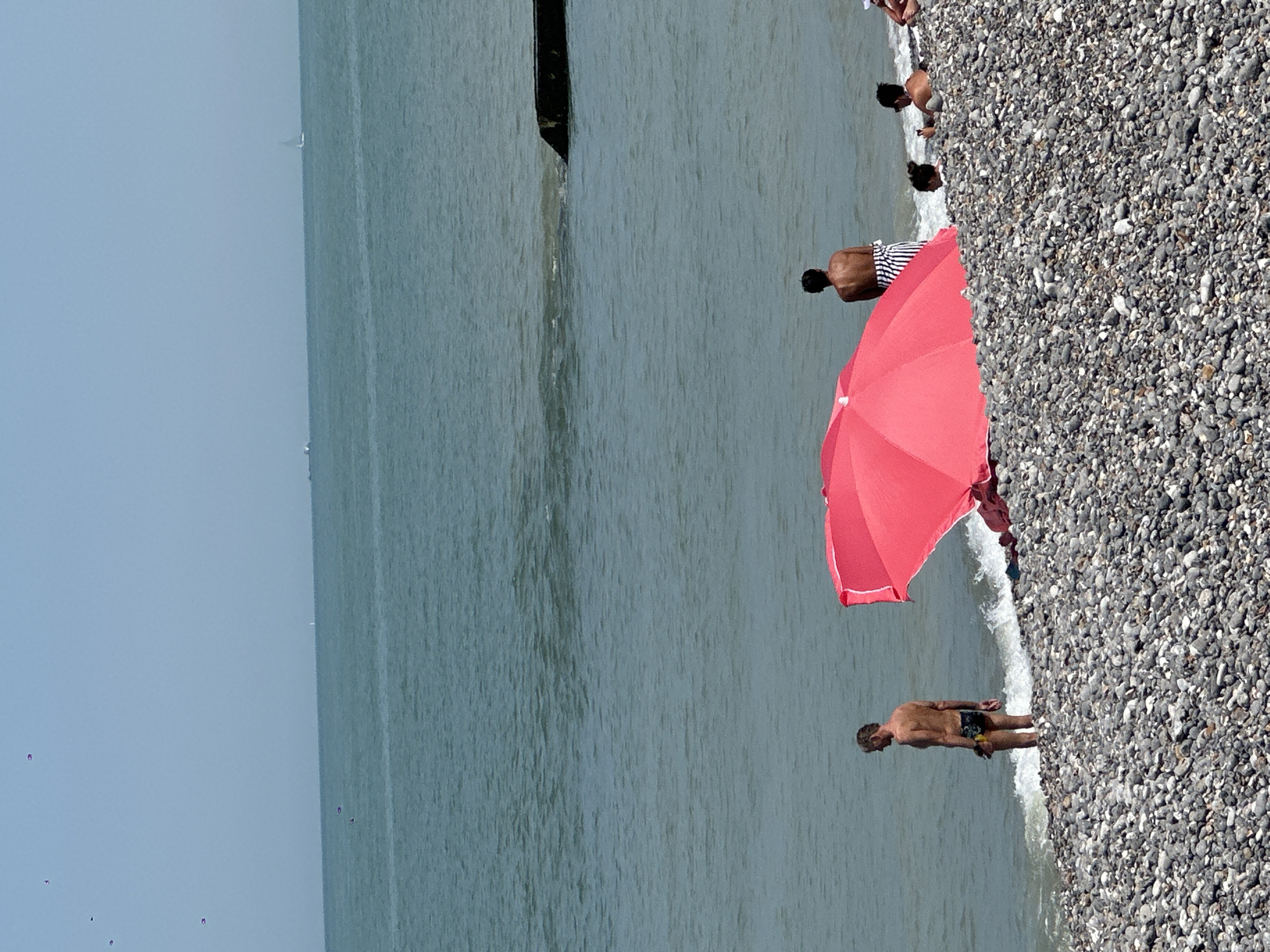 The image depicts a pebble beach in Hautot-sur-Mer, France. In the foreground, a large, open pink umbrella is prominently displayed. To the left of the umbrella, a man in multi-colored swim trunks stands at the water's edge. To the right of the umbrella, a person with their back to the camera, wearing a black and white striped towel or sarong, stands near the water. Further to the right, two individuals are partially visible in the water, with one appearing to hold a phone. In the distance, several small boats and a sailboat are visible on the calm, green ocean. The sky is a clear, pale blue.