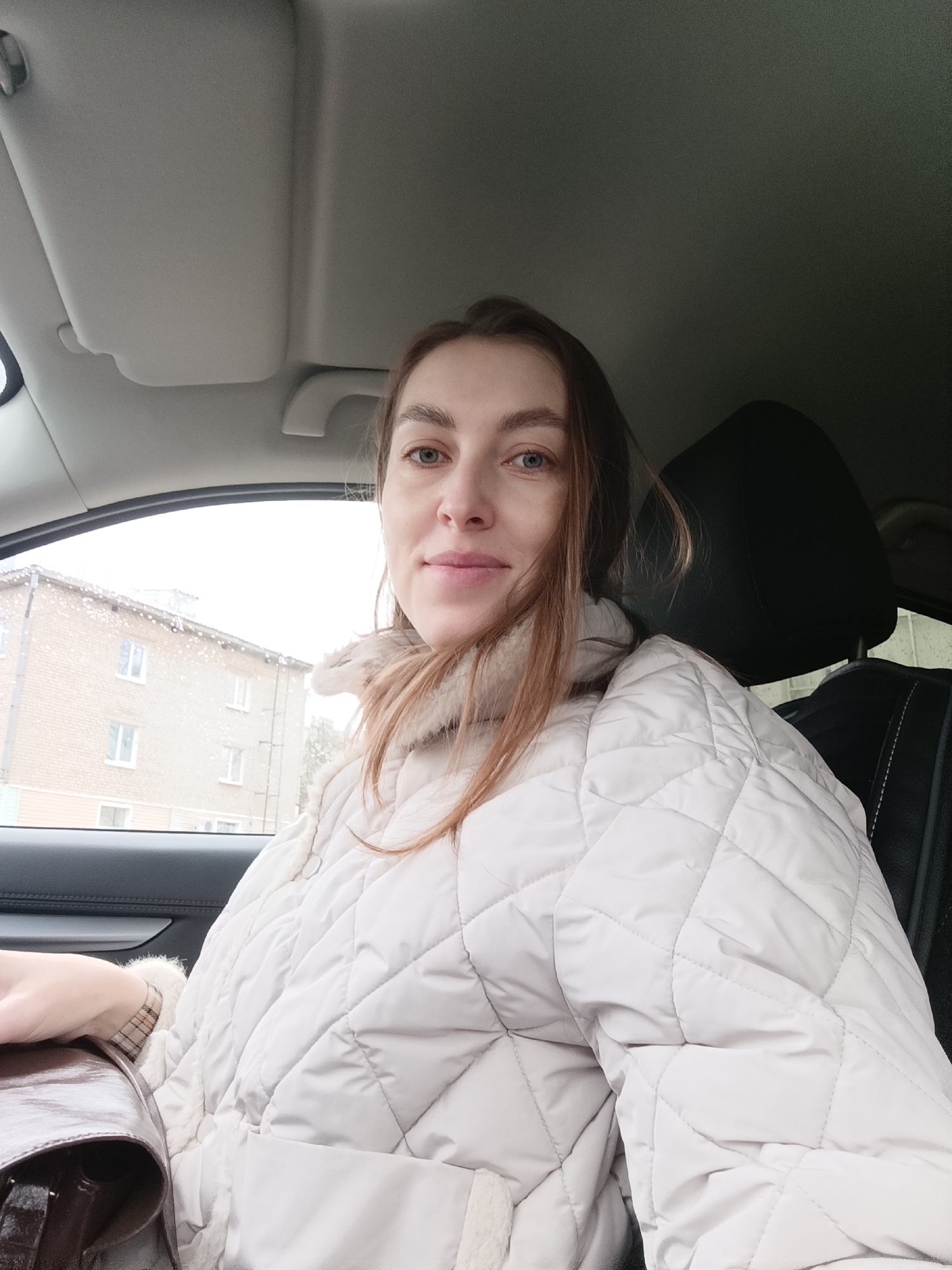 A woman is seated inside a car. The car appears to be in Chaikovsky, Russia, based on the view through the front window of a light brick building with a flat roof and several windows. The weather appears to be overcast, potentially with rain or snow. The woman is fair-skinned, with light brown hair and blue eyes. She is wearing a light-colored, quilted winter coat with a fluffy collar. A dark-colored purse is on her lap or beside her. The car interior is visible, including the headrest of the car seat and the car's roof. The woman is looking directly at the camera, her facial expression appearing neutral.
