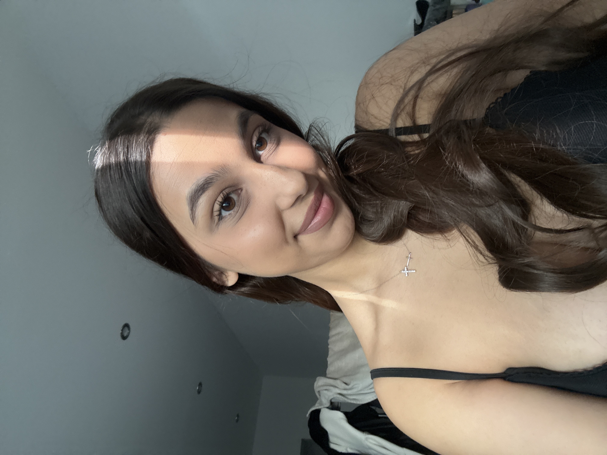 A young woman poses for a selfie. She has long brown hair, brown eyes, and fair skin. She is wearing a black top and a silver necklace with a cross pendant. The background appears to be a bedroom with a white wall and a sloped ceiling. The image does not provide sufficient information to identify the location as Mönchengladbach, Germany.