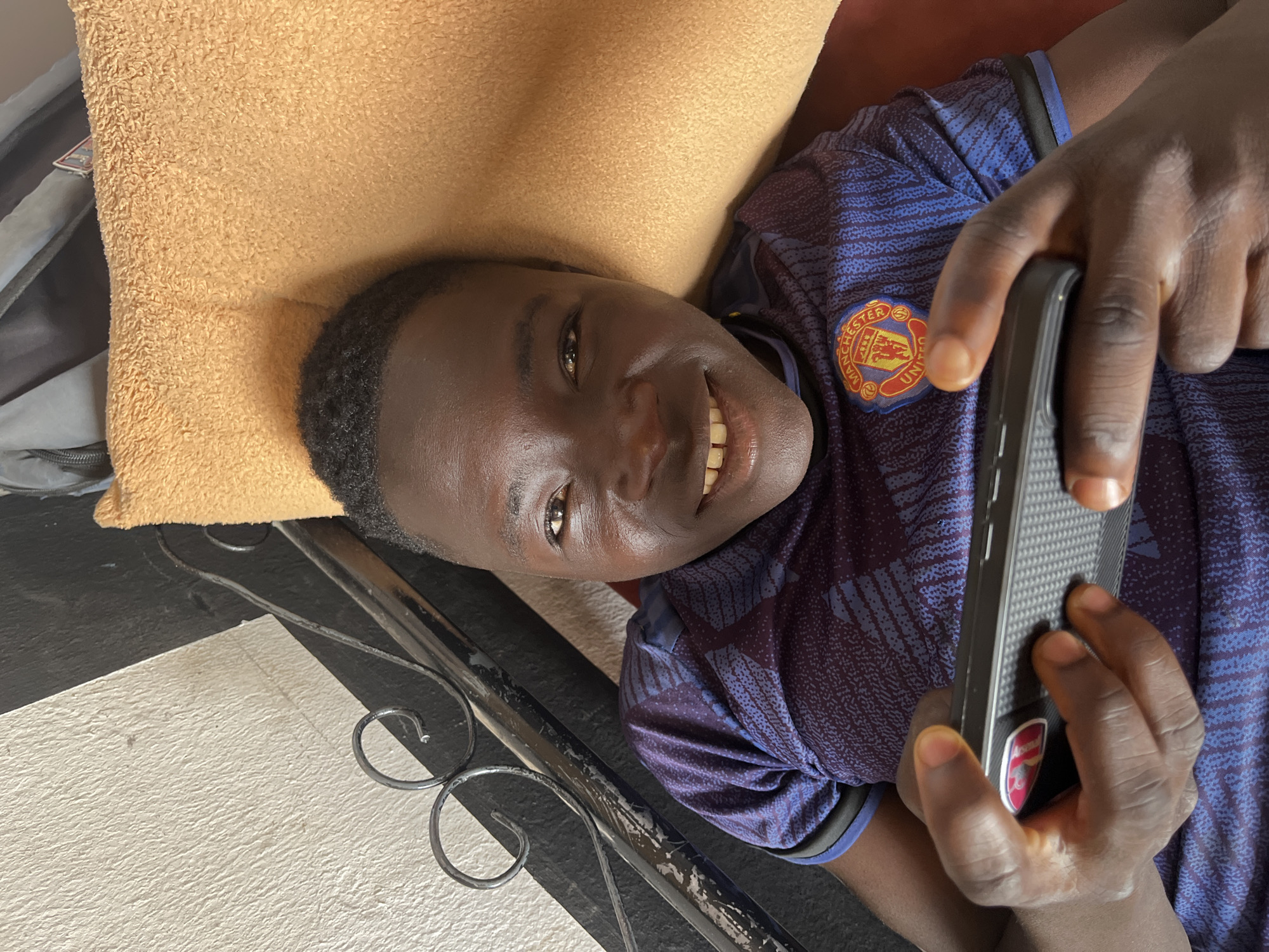 A young, dark-skinned male, likely in his late teens, is lying on a tan pillow, appearing relaxed and smiling at the camera. He's wearing a blue soccer jersey with a Manchester United logo prominently displayed. He is holding a black phone. The background features an ornate black metal railing. The location is potentially Jos, Nigeria, however, there is no evidence in the image to directly confirm this. The setting appears to be indoors. The young man is interacting with the phone, possibly playing a game or browsing.