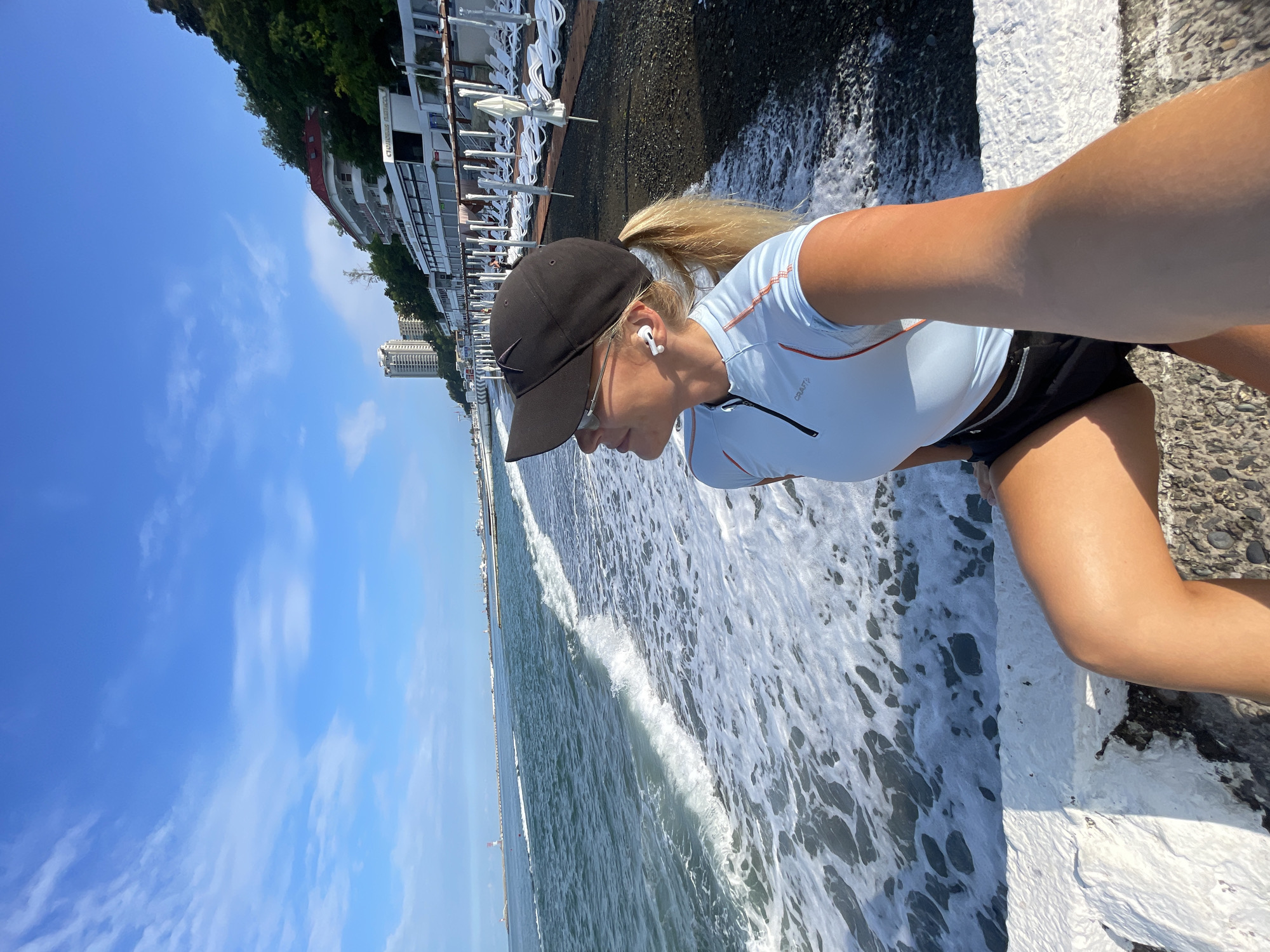 A woman with blonde hair pulled into a ponytail is sitting on a white concrete barrier on a pebble beach in Sochi, Russia. She is wearing a light blue athletic shirt, black shorts, a black baseball cap, and sunglasses. She has white wireless earbuds in her ears and is looking out at the sea. The waves are breaking on the shore, and there are lounge chairs and umbrellas set up on the beach behind her. In the background, there are several buildings and a lighthouse in the distance. The sky is blue with some white clouds.