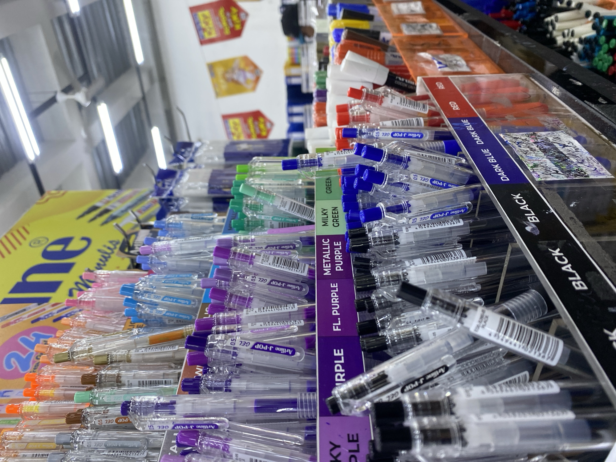 A close-up, eye-level, medium shot captures a display of colorful gel pens in a retail store, likely in Depok, Indonesia. The pens are neatly arranged in clear plastic holders, organized by color and type, with labels such as "BLACK", "DARK BLUE", "RED", "PURPLE", "FL. PURPLE", "METALLIC PURPLE", and "MILKY GREEN" visible. The pens themselves have clear barrels with colored ink cartridges and caps, showcasing a variety of vibrant hues. In the background, fluorescent lights illuminate the store's ceiling, and promotional posters with Indonesian text are displayed on the wall. The overall impression is one of a well-stocked stationery section, showcasing a wide selection of writing instruments.