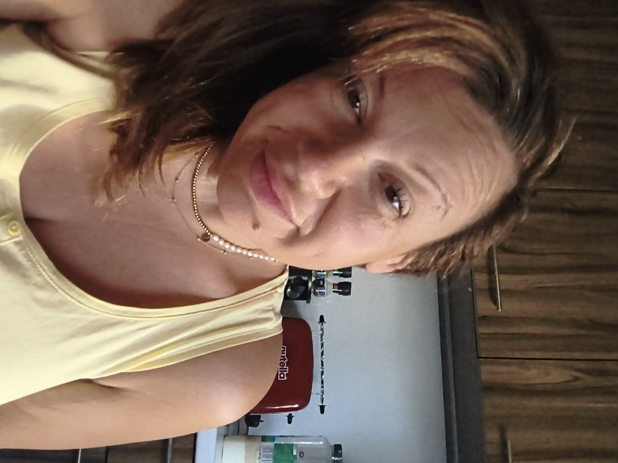 A woman is posing for a photo in what appears to be a kitchen. She is wearing a yellow tank top and a pearl necklace. Behind her is a kitchen counter with a red Nutella toaster oven and some bottles. The background includes a dark wooden cabinet. There is no information available in the image to determine that the location is in Sofia, Bulgaria.