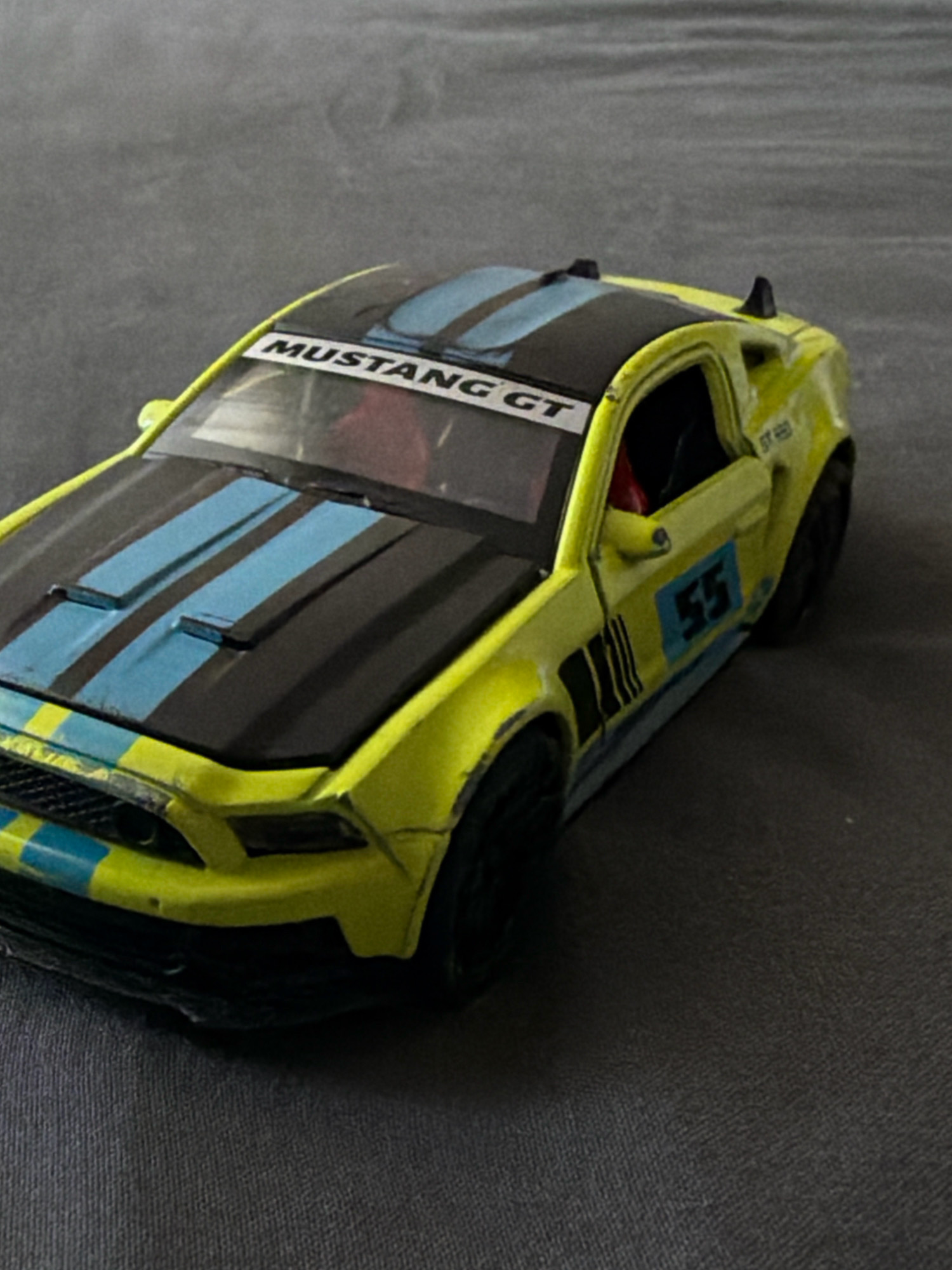 A yellow toy Ford Mustang is displayed on a dark gray textured surface. The toy car features black racing stripes on the hood and blue stripes on the front grille. A "MUSTANG GT" decal is visible on the windshield. The car has racing numbers "55" on the side and "GT 469" on the rear fender. The background is out of focus, suggesting the image was taken indoors. There are no people or any indication of the city of Hrodna, Belarus.