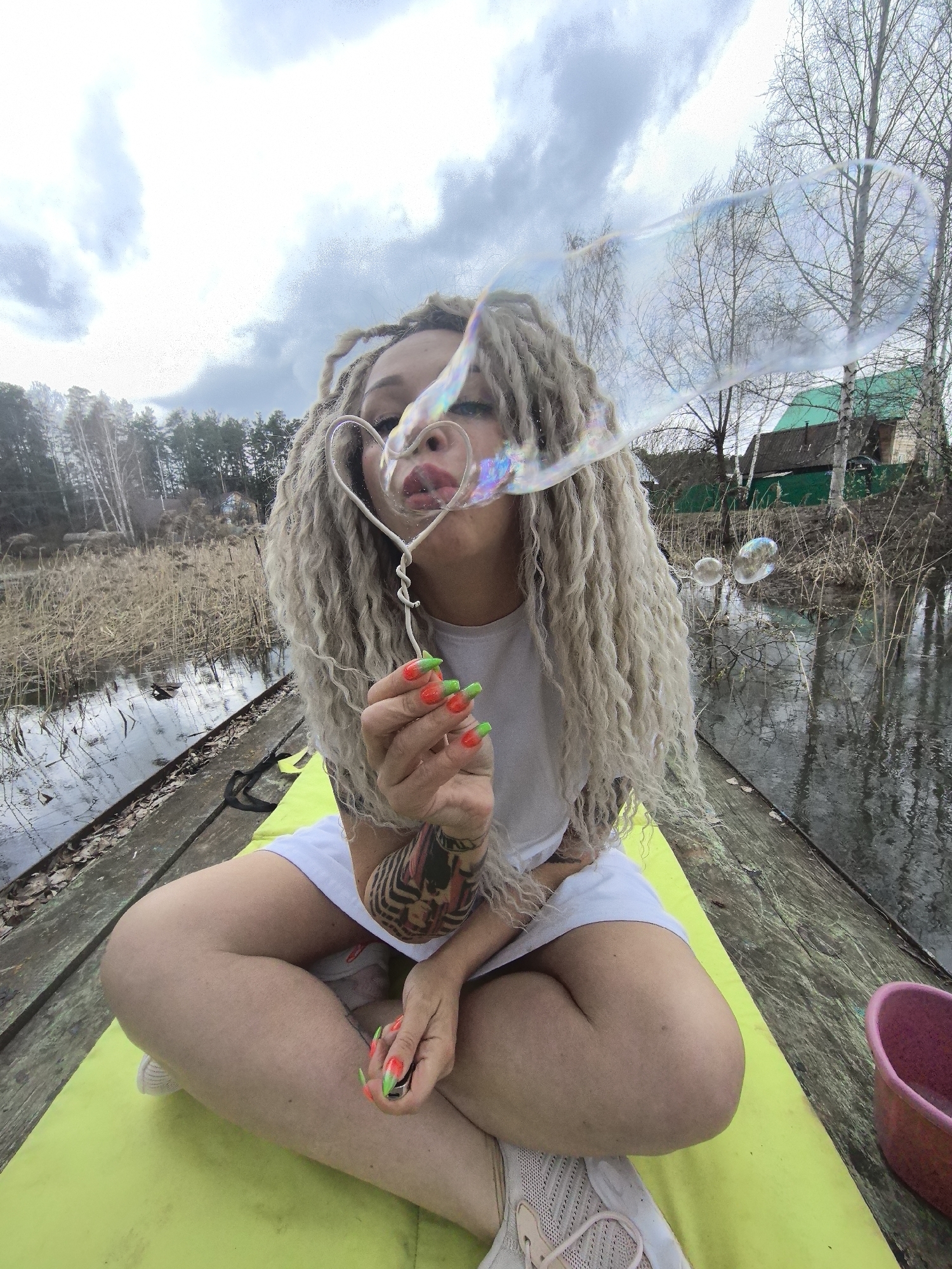 A person is seated cross-legged on a lime green mat on a rustic wooden structure, possibly a boat or a small pier, at the edge of a body of water. The subject has long, blonde dreadlocks or tightly coiled curly hair and is wearing a white t-shirt and white shorts or a skirt. A tattoo is visible on their right forearm. Their fingernails are brightly painted with a gradient of neon orange and green.
The central activity is the person blowing soap bubbles. They hold a heart-shaped bubble wand to their lips, from which a large, elongated, iridescent bubble is emerging towards the left. Several smaller, spherical bubbles are seen floating over the water to the right, reflecting the cloudy sky.
The scene is outdoors during the daytime, under a cloudy, overcast sky. The surrounding environment suggests a rural or semi-rural setting, possibly in Noviy, Russia, during early spring or late autumn, given the bare deciduous trees. The body of water, likely a lake or a calm river, is flanked by tall, dry reeds or grass. In the background to the left, a dense forest with both evergreen and bare trees is visible. To the right, across the water, there are small wooden houses with dark roofs and some green accents, nestled among more bare trees. A pink bucket or bowl is visible on the wooden surface in the lower right corner. The water reflects the sky and the surrounding bare branches. No visible text is present.