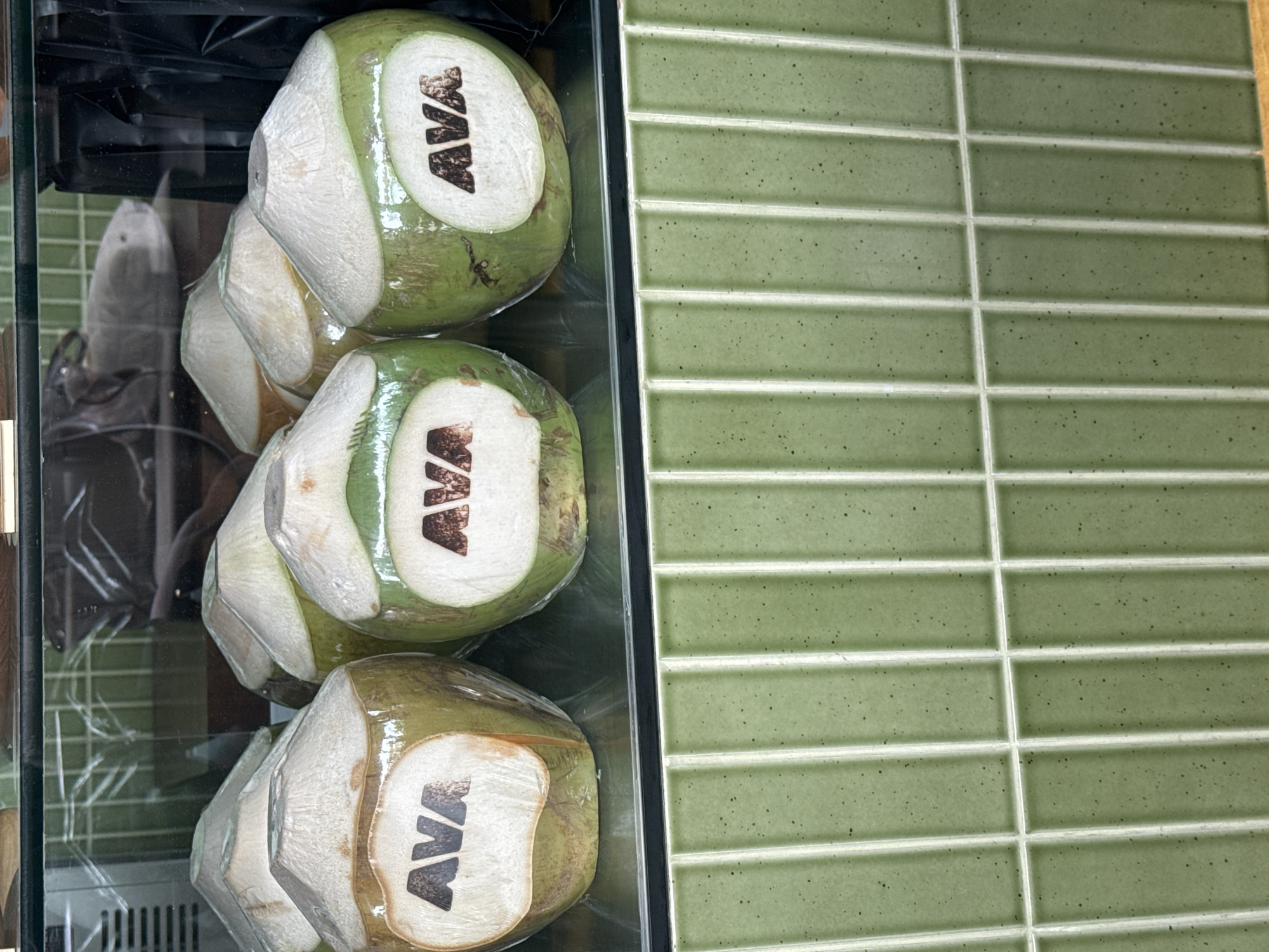 This is a close-up, eye-level, indoor shot of several green coconuts displayed in a refrigerated case with a green tiled background. The coconuts are partially wrapped in clear plastic, and each has a white sticker with the black logo "AVA" on it. The lighting is bright and even, illuminating the coconuts and the reflective surface of the case. The overall impression is one of a clean and well-organized display of fresh produce. The location is not discernible from the image.