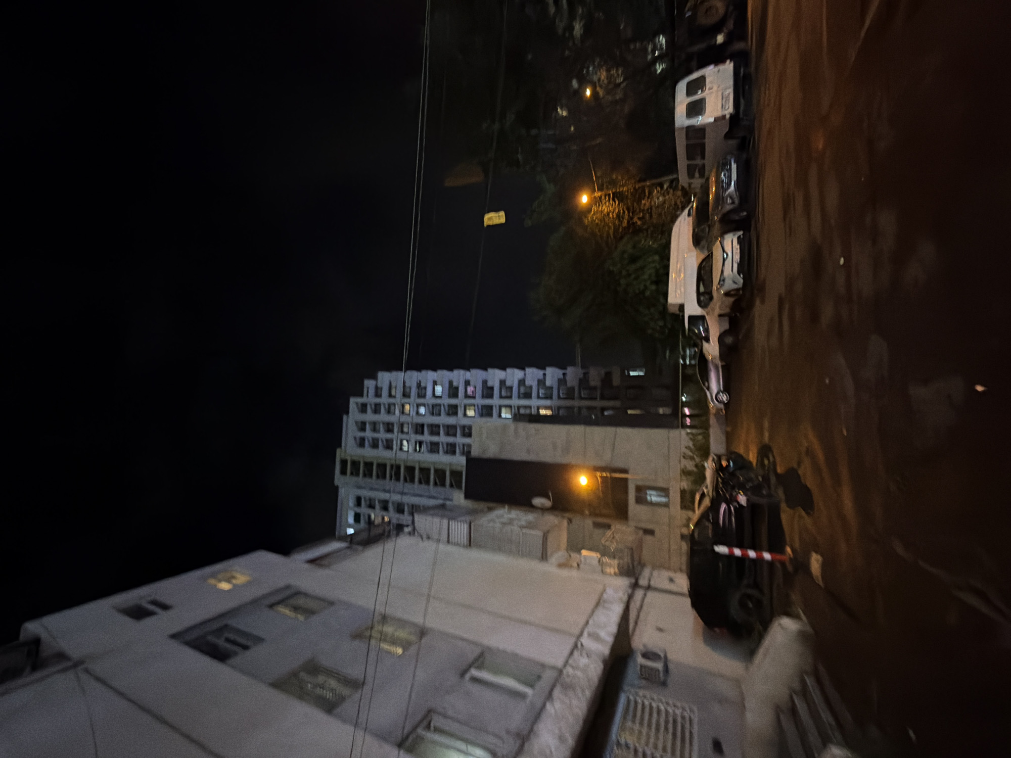 This is a nighttime, low-angle, full-shot photograph taken in Tbilisi, Georgia. The scene depicts an urban environment with residential buildings and parked vehicles.
On the left, a light-colored apartment building, with multiple stories and visible windows, dominates the foreground. Some windows are illuminated, suggesting occupancy. There is an air conditioning unit visible on the side of this building.
In the mid-ground, a dark-colored SUV is parked at an angle, appearing to be close to a concrete structure. A red and white striped traffic cone is positioned near the SUV. Further to the right, several other vehicles are parked, including white vans and sedans. The road surface appears wet, reflecting the ambient light.
In the background, a taller, more modern apartment building with numerous windows is visible, also with illuminated window panes. Trees and foliage are present, particularly on the right side of the frame, and their branches are silhouetted against the dark sky. A streetlamp is illuminated, casting a warm glow. A yellow rectangular object is suspended from overhead wires. The sky is dark, indicating nighttime.