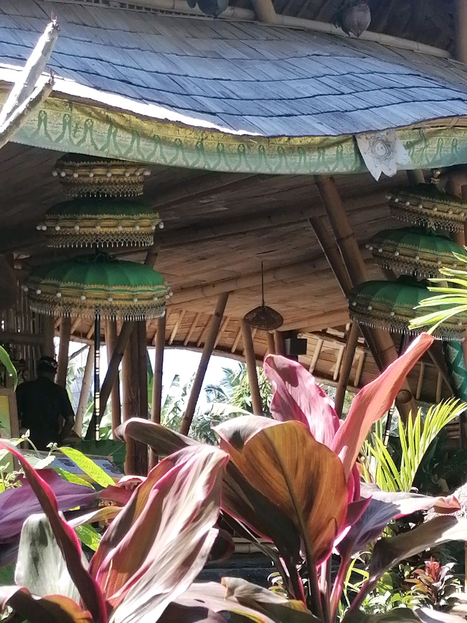 In Ubud District, Indonesia, an outdoor cafe features a thatched roof made of blue-grey tiles, supported by bamboo beams. Ornate, stacked green umbrellas with gold trim and white pearl-like embellishments hang from the ceiling. A man in a black shirt and head covering stands facing away from the camera, looking towards the outdoors. In the foreground, lush green and red foliage with broad leaves dominates the view. The environment appears to be a tropical setting, with sunlight filtering through the structures and vegetation.