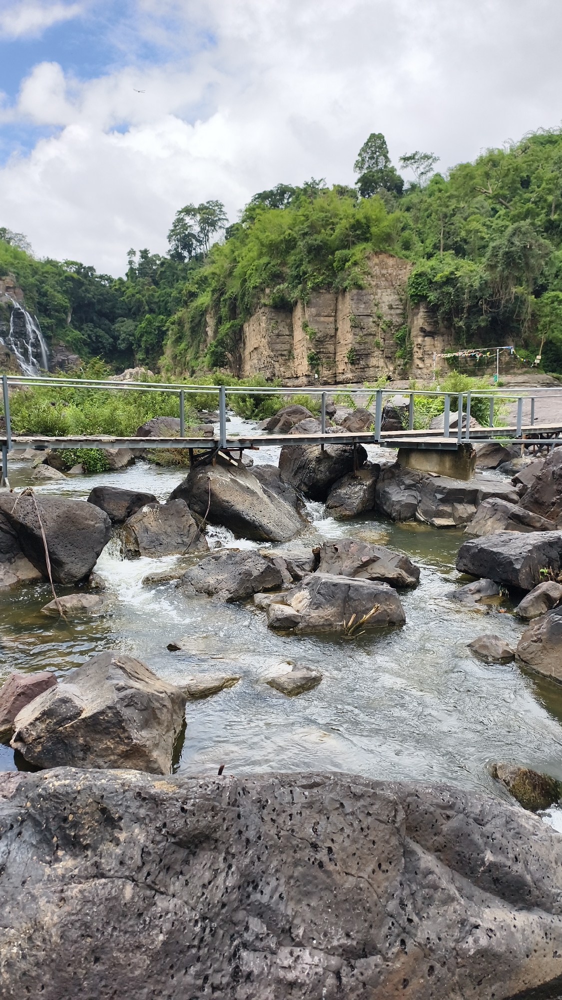 This is a vertical, medium-shot photograph of a rocky river flowing under a metal footbridge. In the background, lush green trees cover a steep, rocky cliff face. To the left, a waterfall cascades down the cliff. The sky above is filled with scattered clouds. The river is filled with large, dark rocks of various sizes, some submerged and others partially exposed. The water flows rapidly around the rocks, creating white foam. The footbridge, made of metal railings and wooden planks, spans the river, connecting the two sides. The scene is set in a natural, outdoor environment. The location is the Suối Tranh waterfall in Tân Phú, Vietnam.
