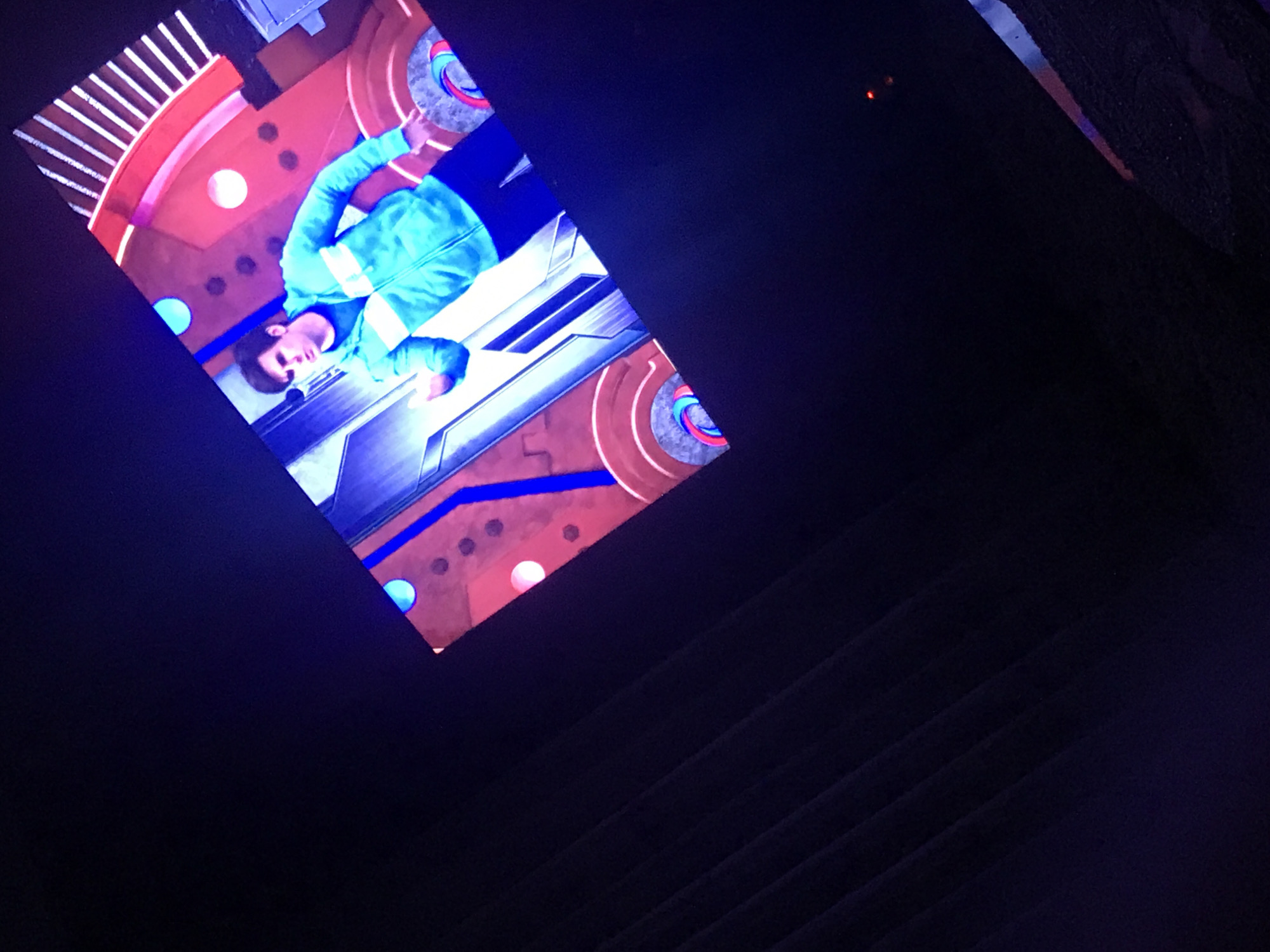 The image captures a scene indoors, likely within a building. Dominating the view is a tilted screen displaying a male figure clad in a light blue jacket with white stripes. He is positioned on a set featuring a mix of architectural elements, including red and white striped patterns and round shapes. A futuristic-looking platform or structure is also visible behind the man. The lower right portion of the image is obscured by a dark, shadowed area, potentially suggesting the presence of steps. The precise location in Yerevan, Armenia, cannot be determined based on the information provided in the image.