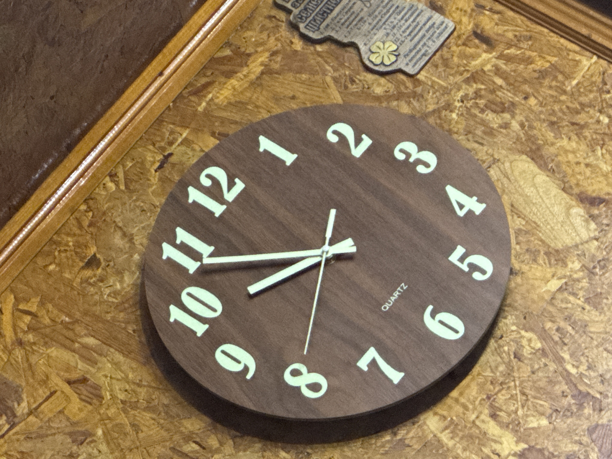The scene is an interior view in Sarnevo, Bulgaria, featuring a circular wall clock mounted on a textured wood paneling surface. The clock has a dark brown face with prominent, luminous light green numerals from 1 to 12. The word "QUARTZ" is printed in white capital letters on the lower left quadrant of the clock face, between the 7 and 6. The clock's three luminous hands indicate the time: the hour hand is precisely aligned with the 8, the minute hand is aligned with the 12, and the thin second hand is aligned with the 8. This indicates the time is 08:00:40. The wall paneling behind the clock is consistent with oriented strand board (OSB), displaying various compressed wood chips in shades of light brown and tan. To the right of the clock, a partially visible wooden decorative plaque, shaped irregularly, is affixed to the OSB. This plaque features a four-leaf clover symbol and includes discernible Cyrillic text, containing the word "счастье" (happiness). A light-colored wooden frame with a grooved profile runs diagonally across the upper right and lower right sections of the image.