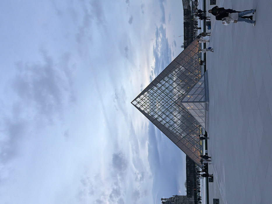 The Louvre Pyramid stands tall against a cloudy sky at dusk. The geometric glass structure reflects the soft light, contrasting with the ornate building below. People stroll across the plaza around the pyramid, their figures appearing small against the vast space. The scene captures the modern design juxtaposed with classical architecture.